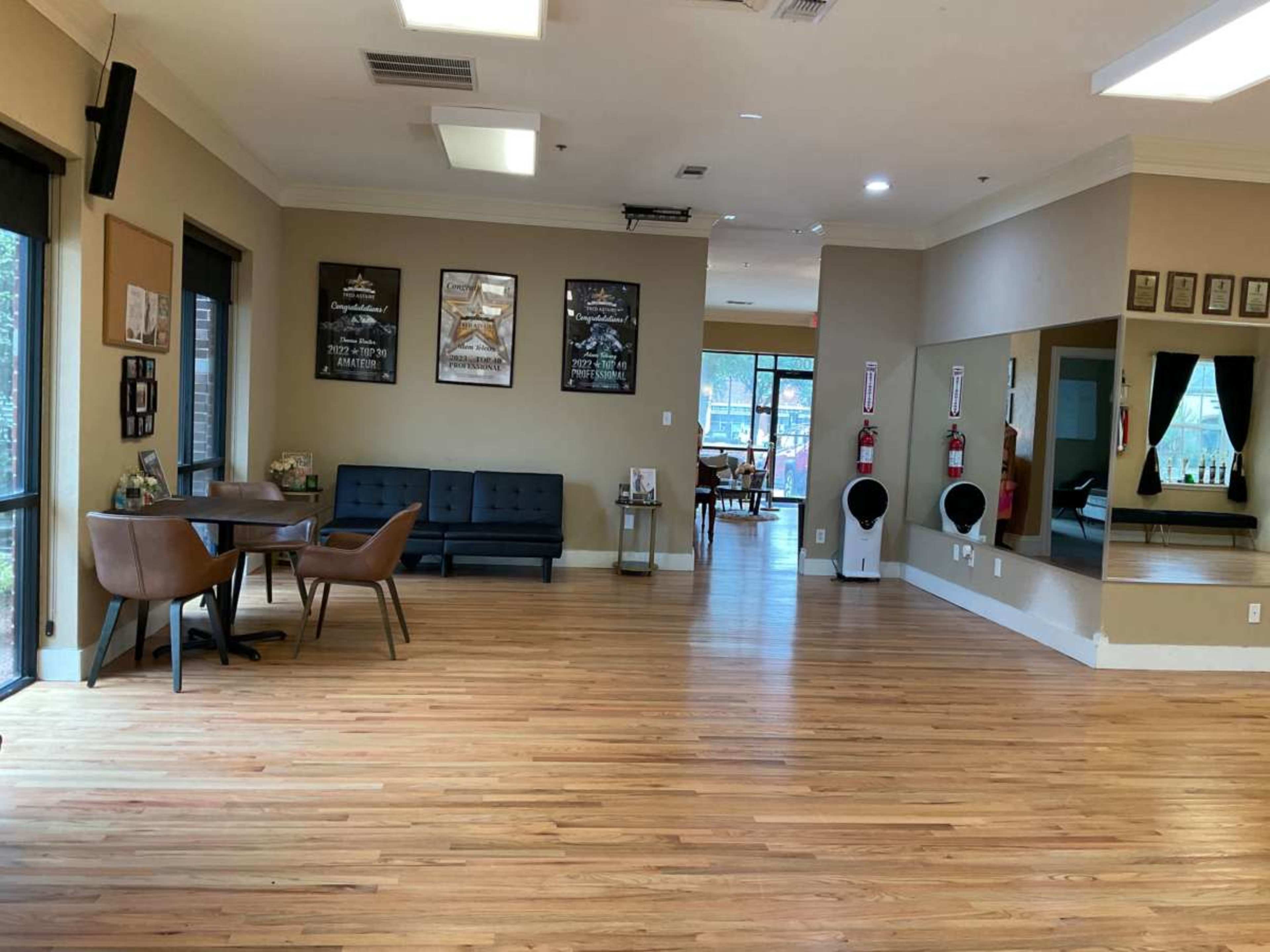 The image shows a spacious interior of a studio or lounge area with wooden flooring, featuring a seating area with chairs and a table, and large windows allowing natural light to enter.