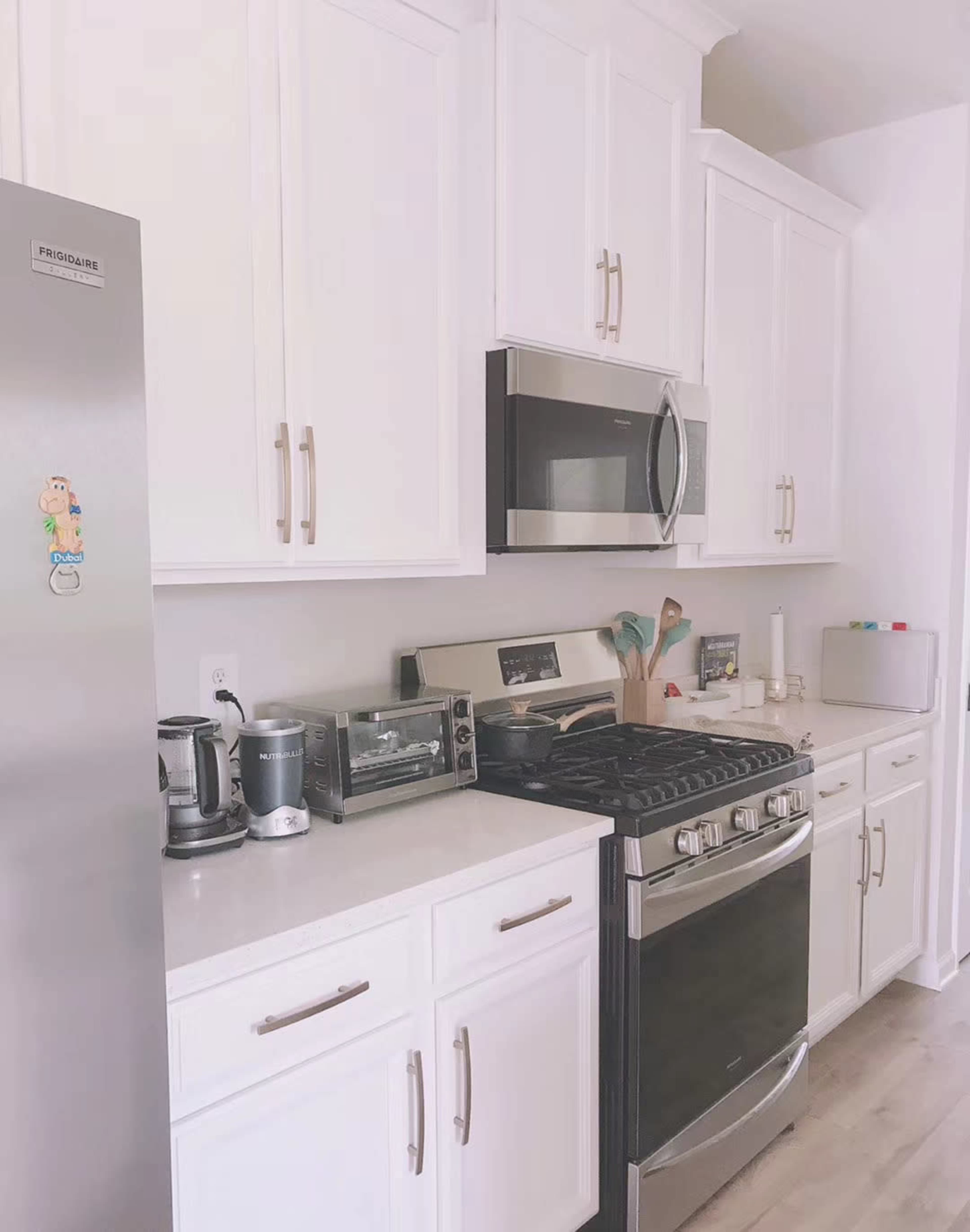 The image shows a modern kitchen with white cabinets, stainless steel appliances, and a countertop lined with various kitchen utensils.