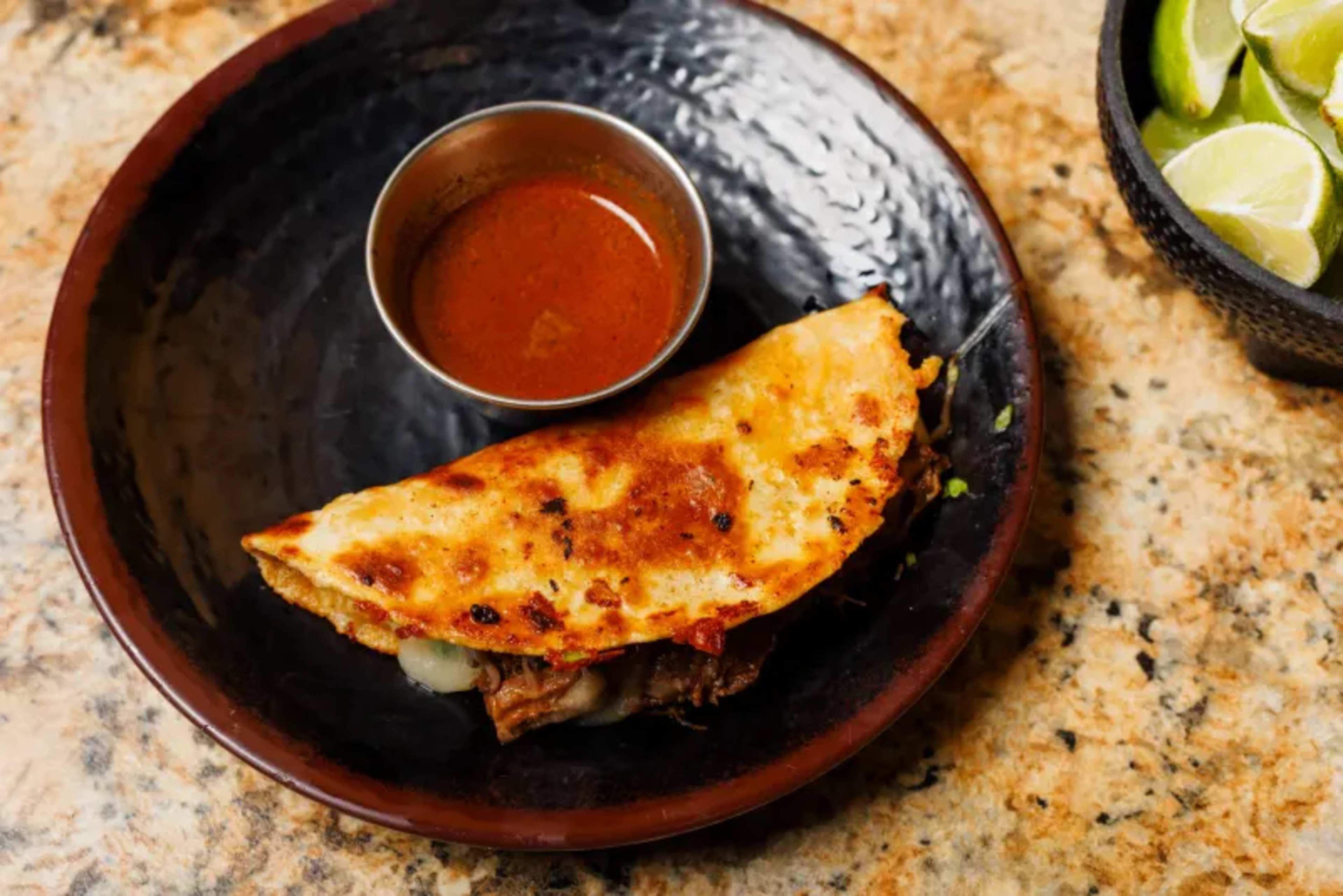 A golden-brown quesadilla rests on a dark plate alongside a small bowl of red salsa and a separate bowl of lime wedges.