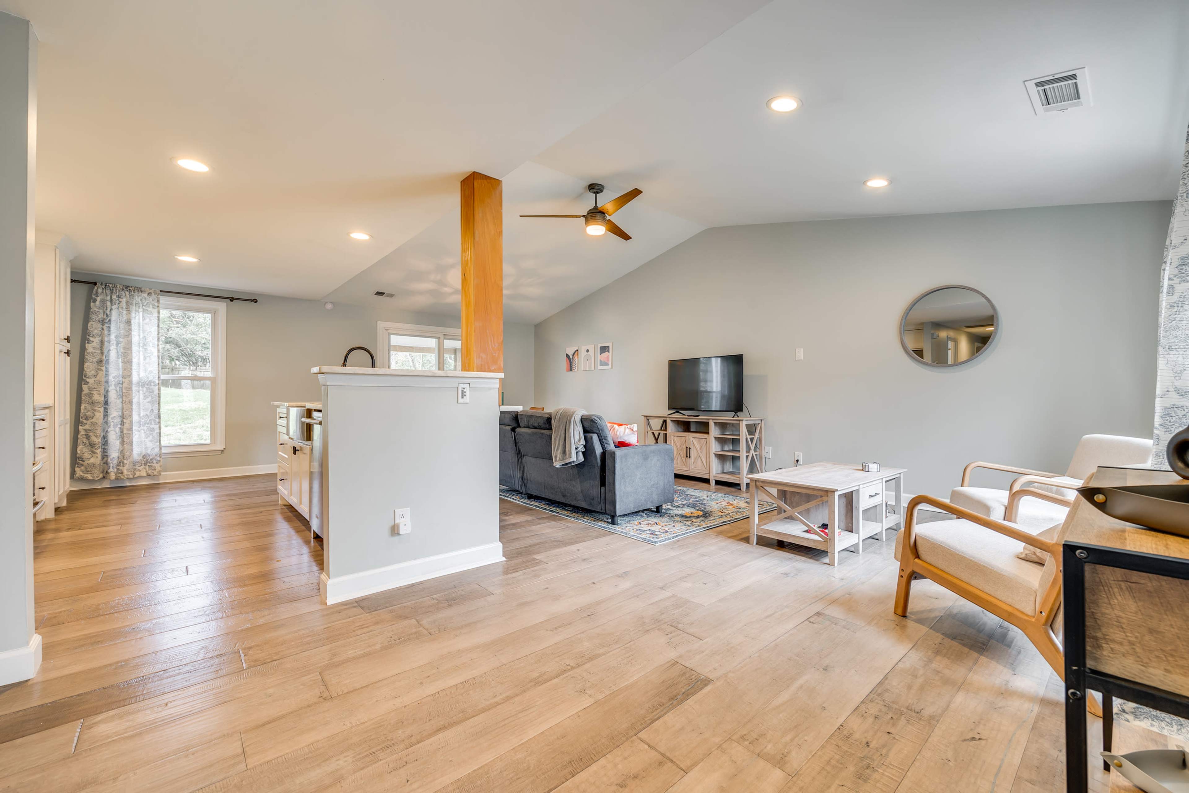 A spacious living area with a gray sofa, wooden flooring, and a ceiling fan, adjacent to a kitchen area with a large window and bright lighting.
