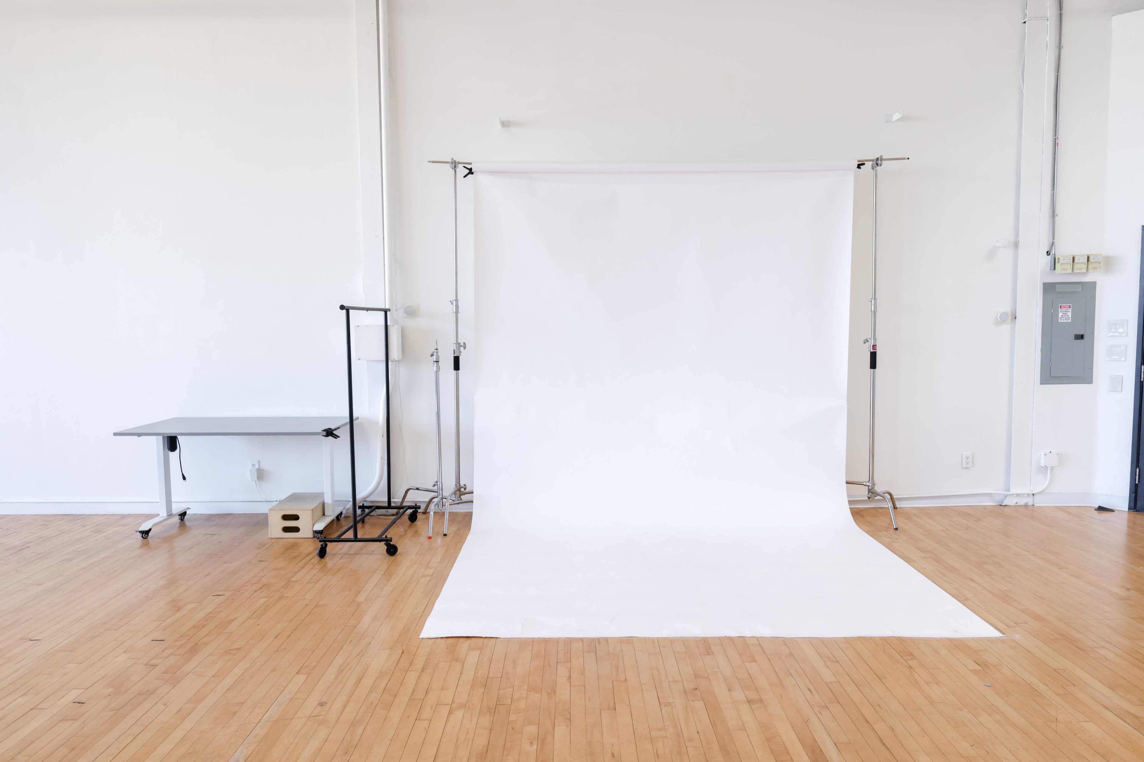 The image shows an empty studio space featuring a white backdrop on stands, a wooden floor, and minimal furniture including a table and a clothing rack.