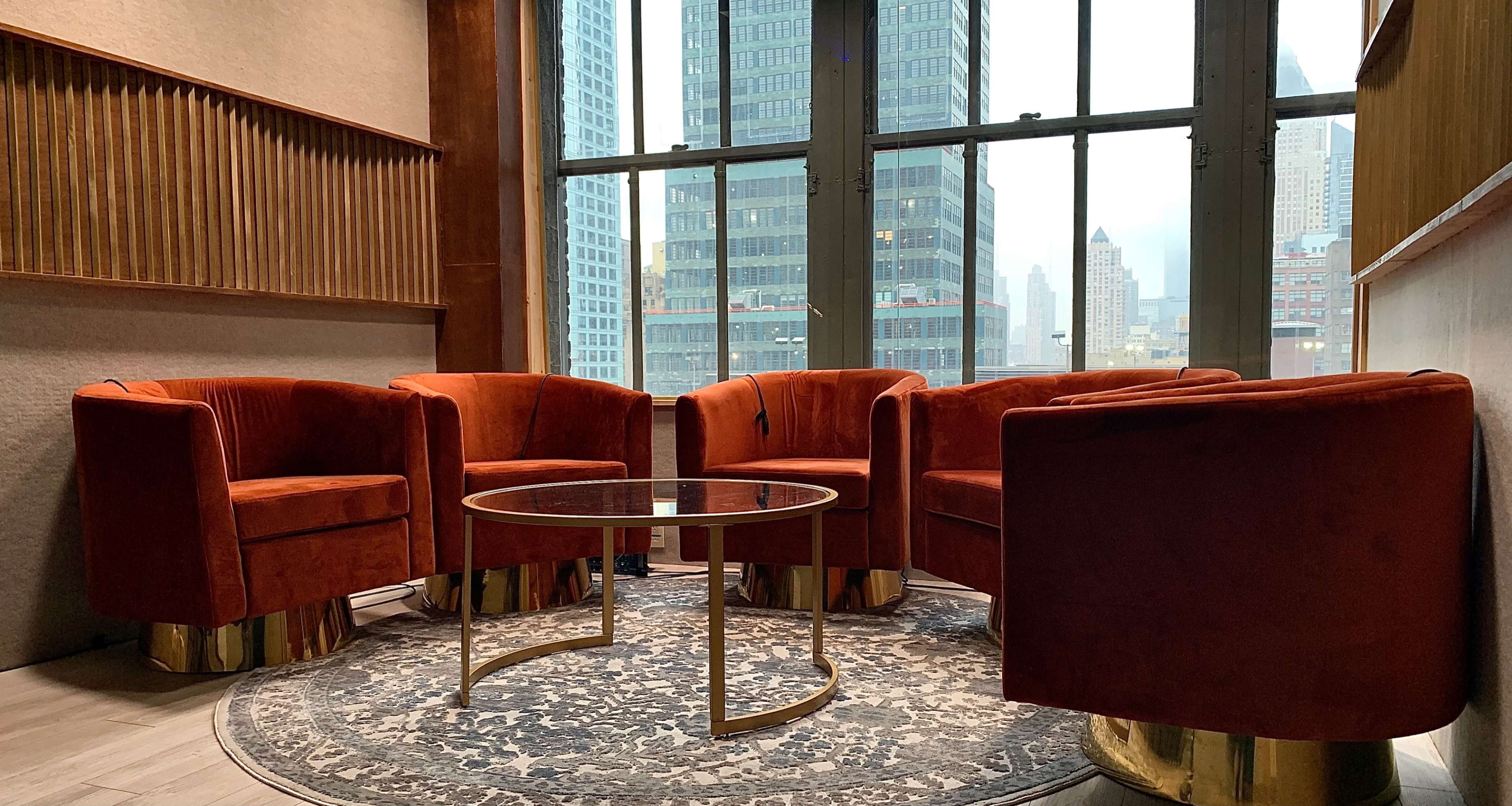 Four orange velvet chairs arranged around a round glass table on a patterned rug, with large windows displaying a city skyline in the background.