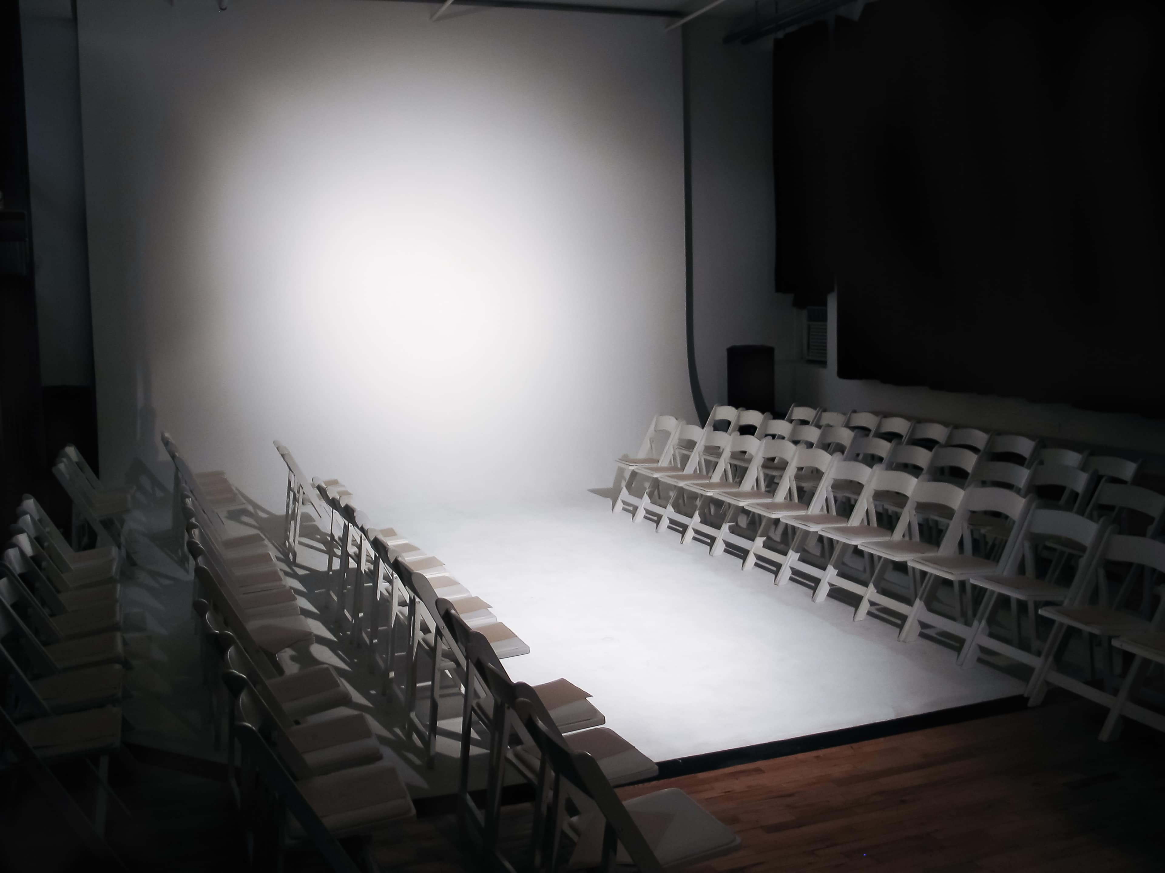 A row of white folding chairs is arranged in two parallel lines facing a brightly lit stage in an empty room.
