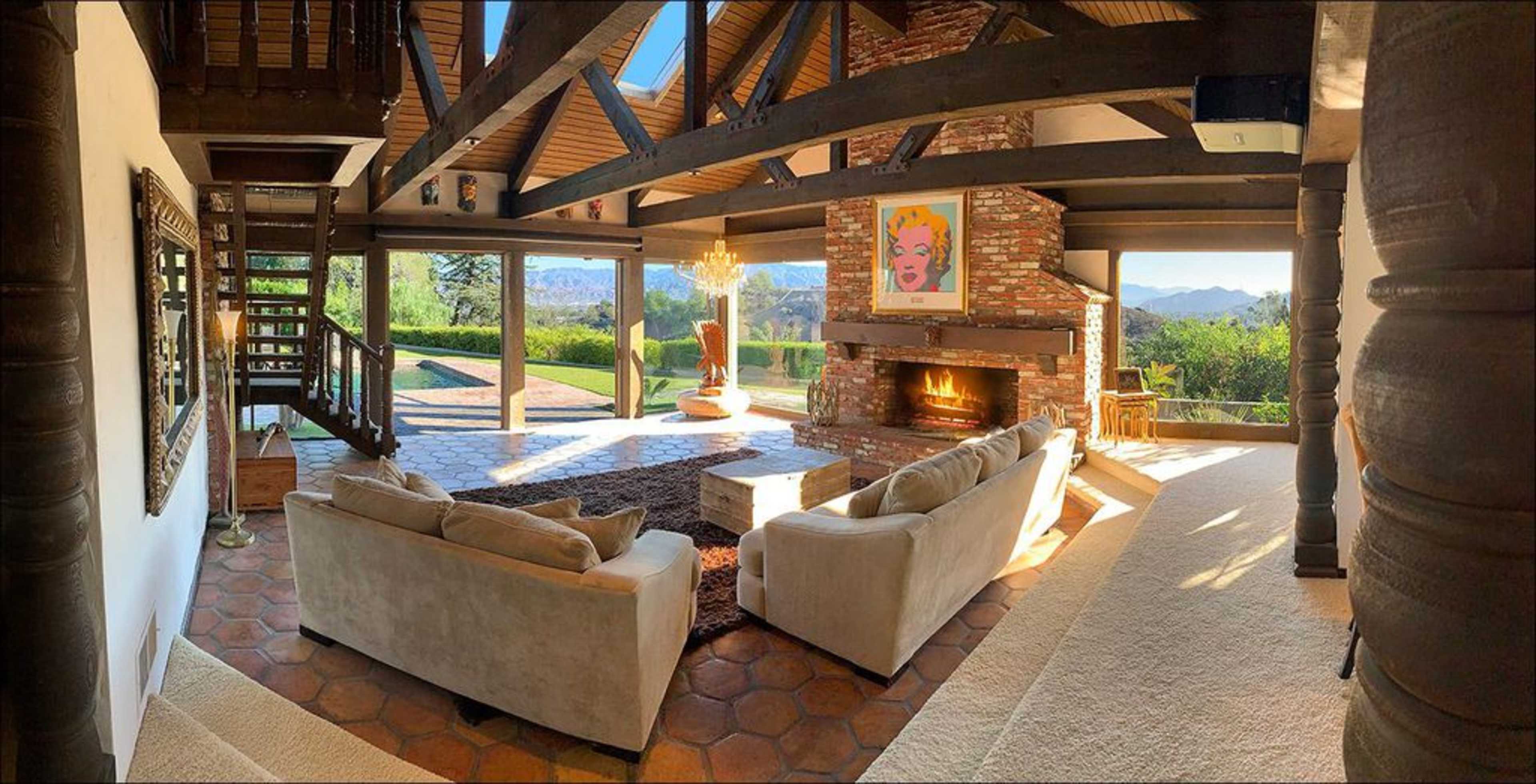 A spacious living room with a stone fireplace, large windows overlooking a landscape, and modern furniture arranged around a central coffee table.