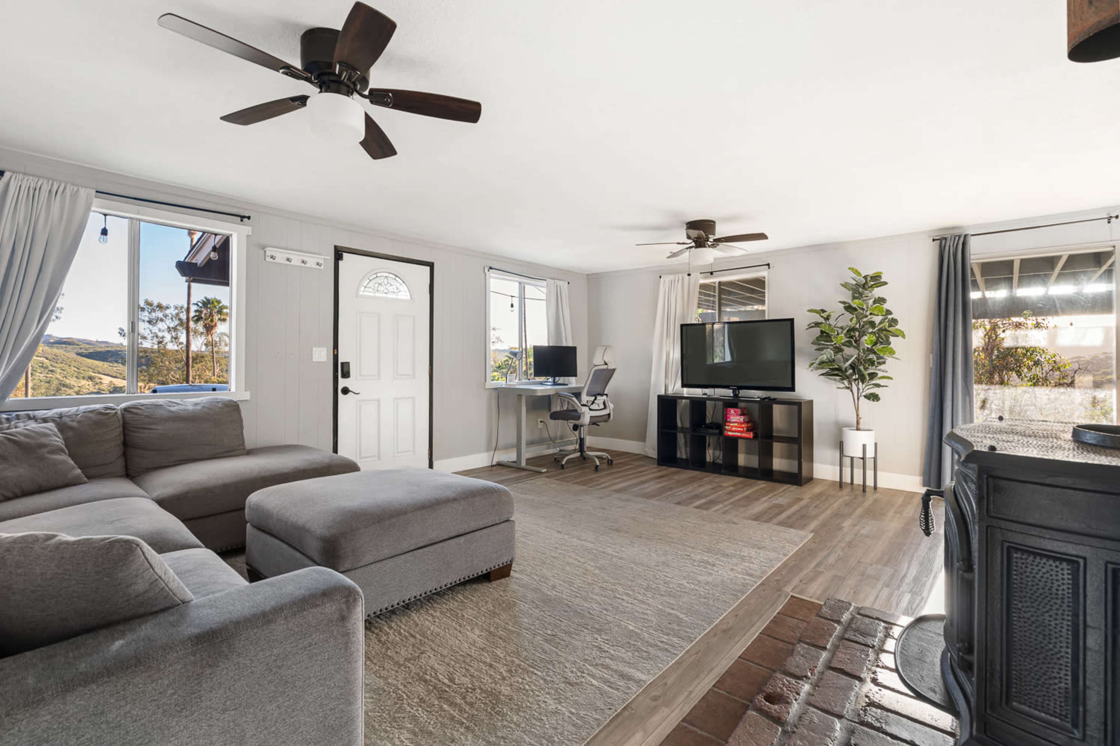 The image shows a living room with a gray sectional sofa, a television on a black stand, a desk with a computer, and large windows allowing natural light to enter.