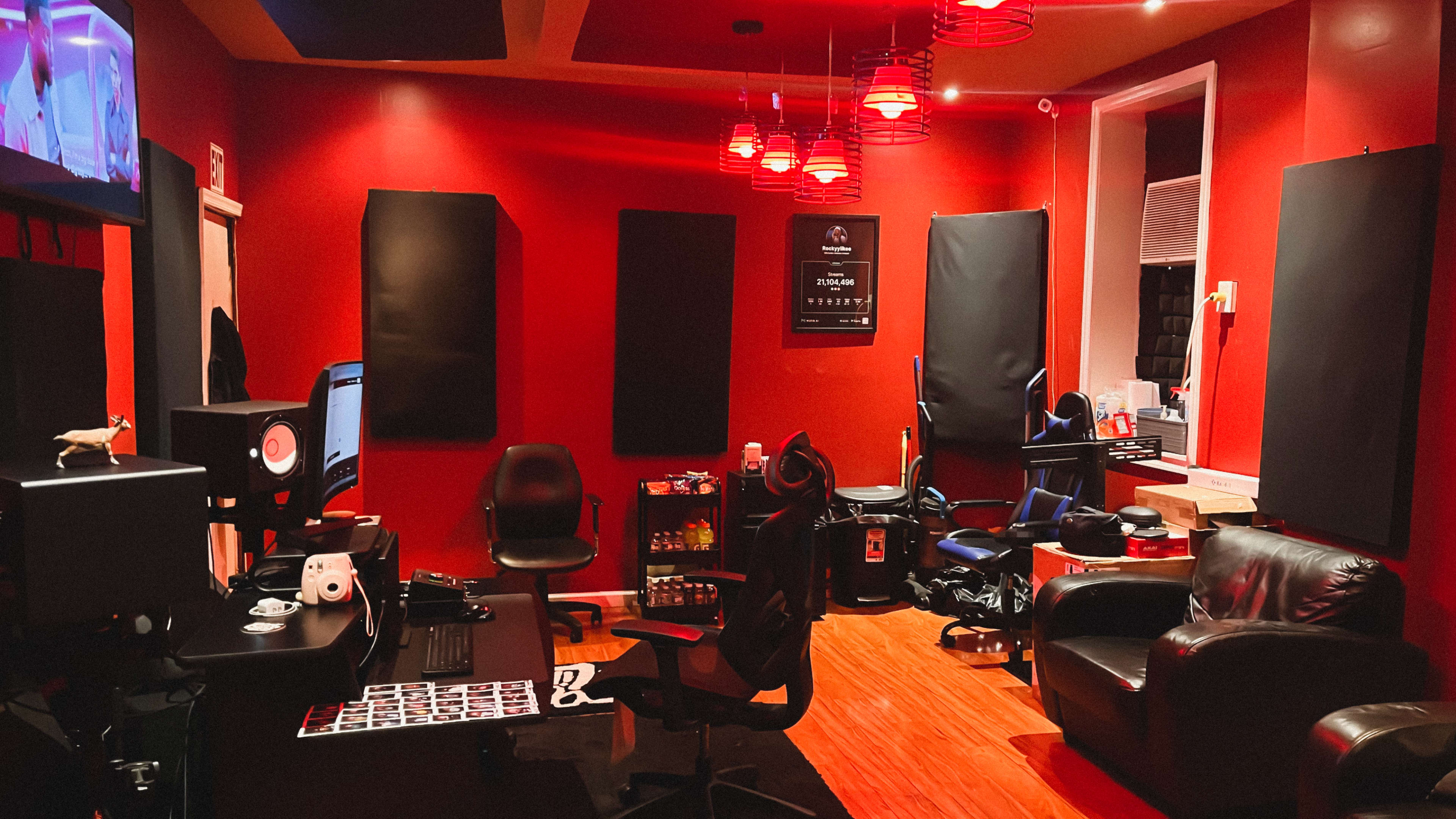 The image shows a recording studio with red walls, black soundproofing panels, multiple chairs, and a desk equipped with music production equipment.