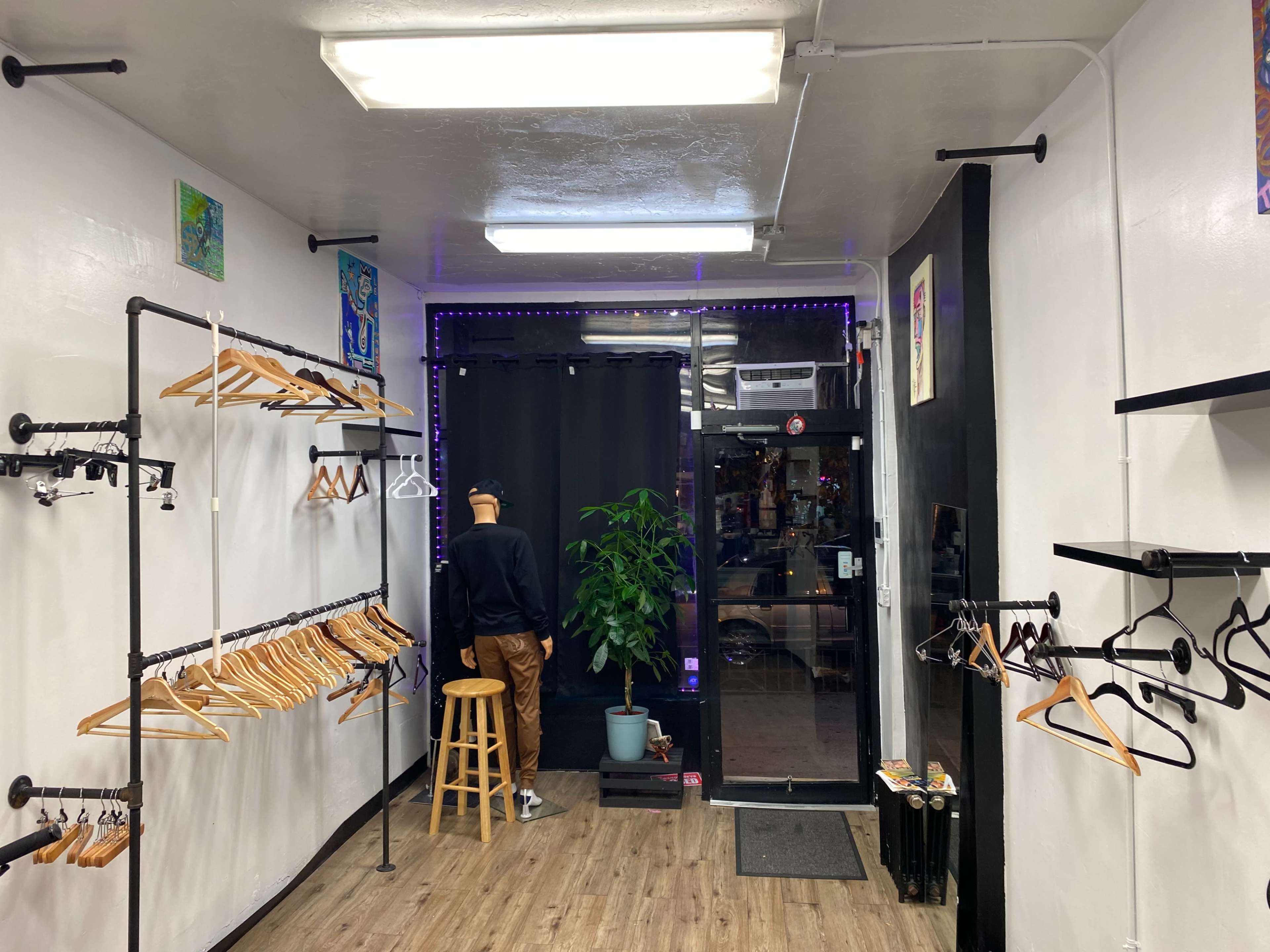 A clothing store interior with wooden hangers on metal racks, a stool, a plant in a pot, and a mannequin facing a curtained entrance.