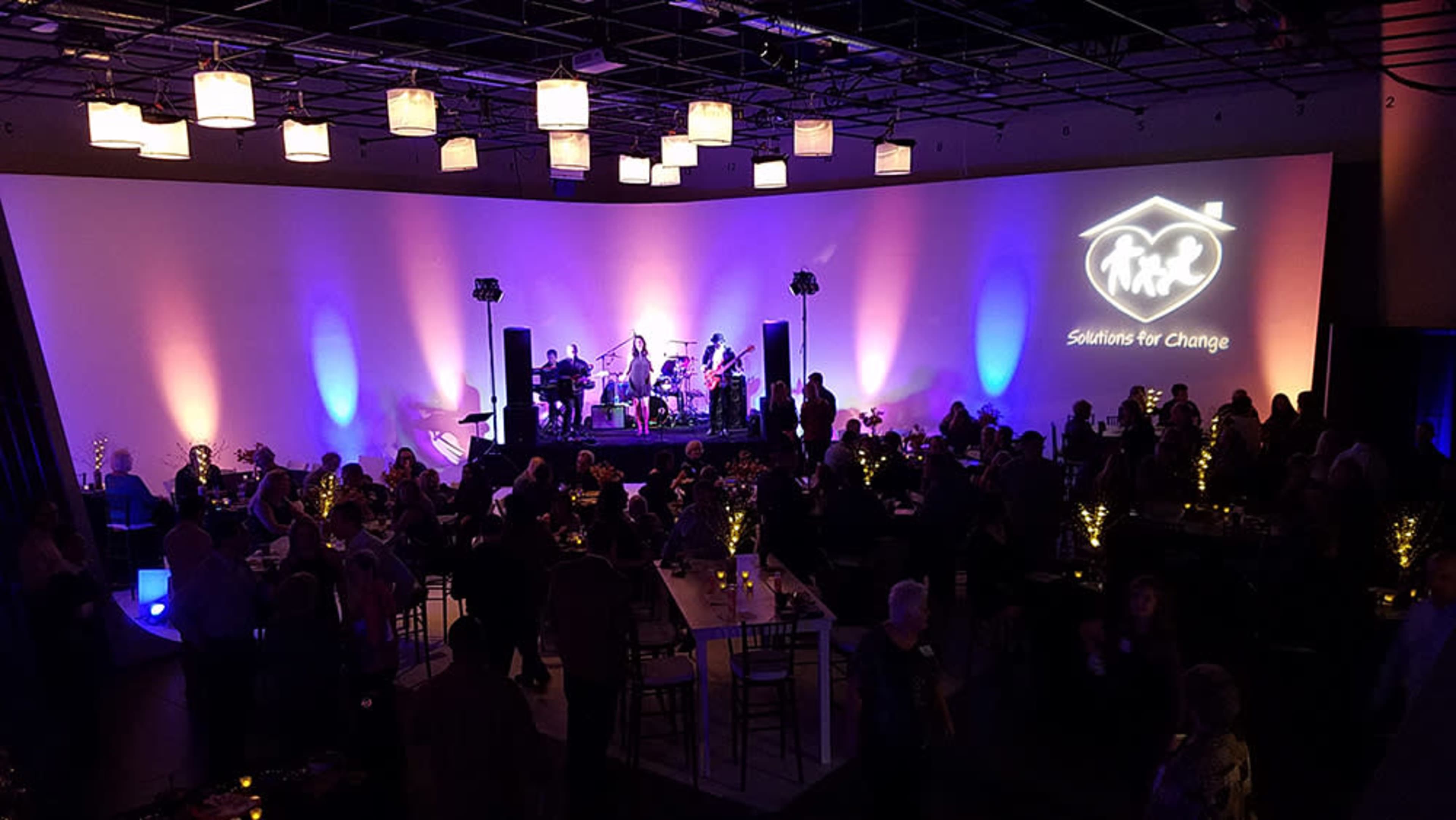 A crowd gathers in a spacious venue with a band performing on stage, illuminated by colorful lights and featuring a projection on the wall.