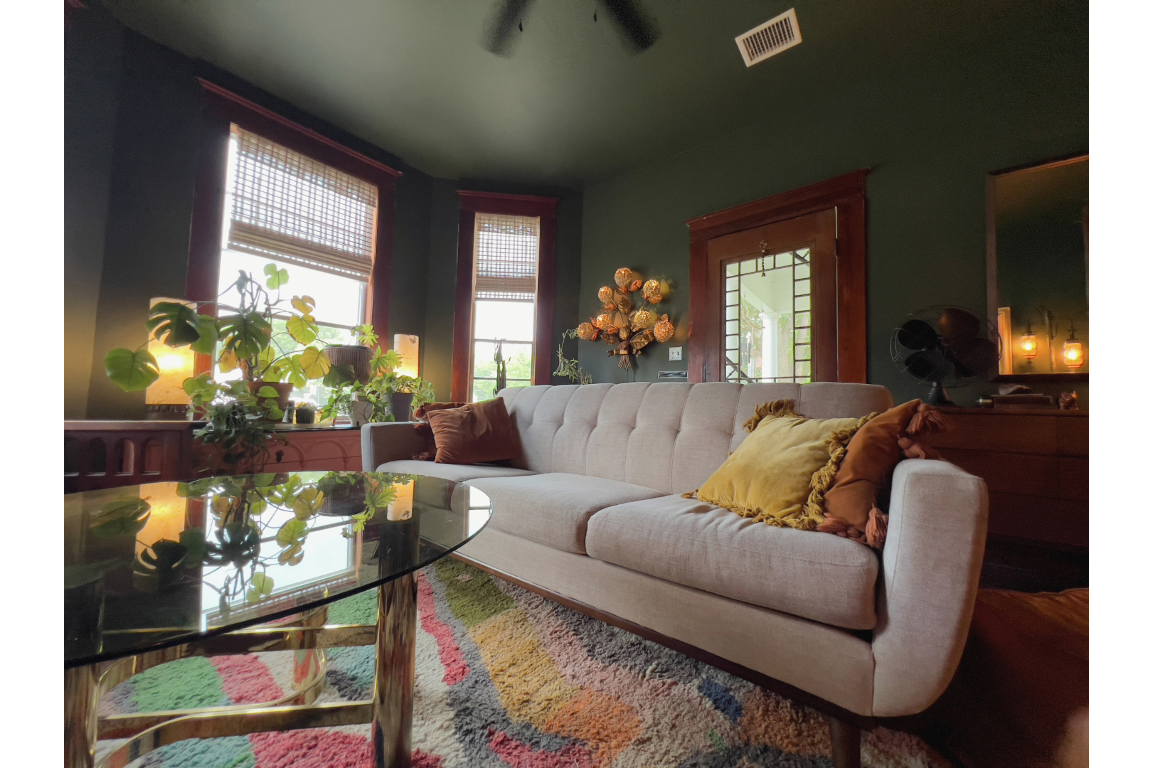 The image shows a cozy living room with a gray sofa, a round glass coffee table, and several plants by the windows, illuminated by soft lighting.
