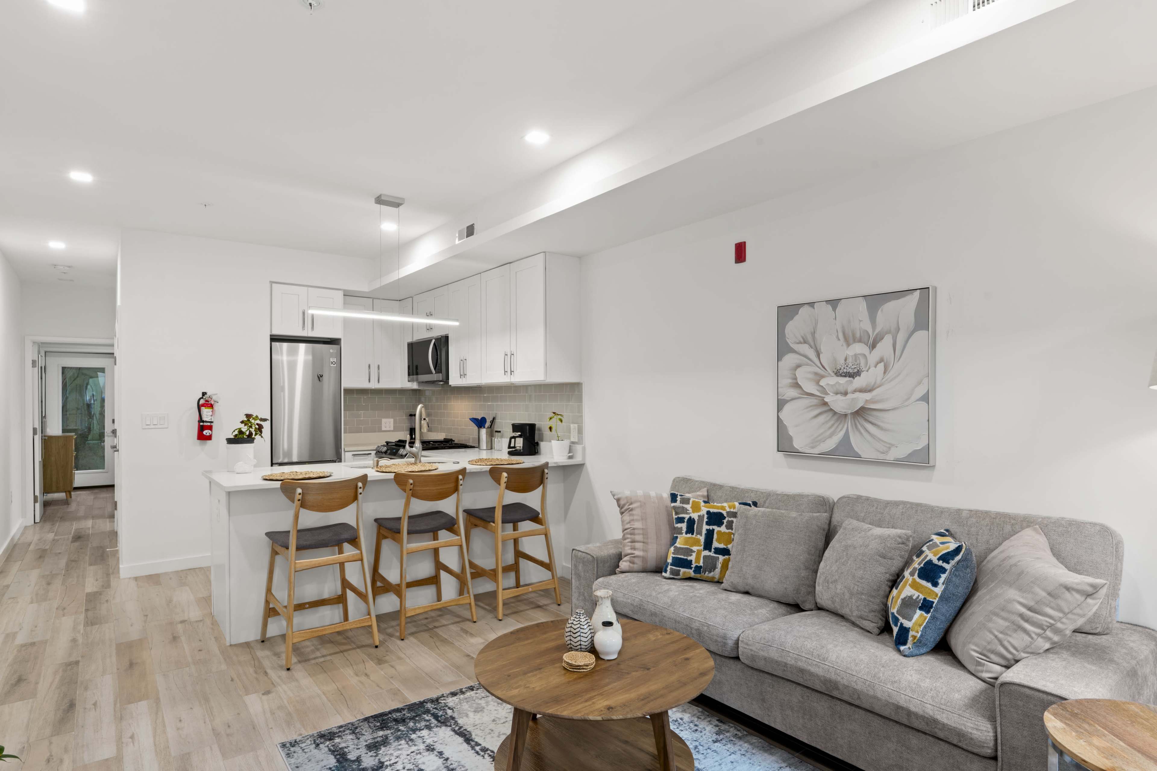 The image shows a modern living area featuring a gray sofa, a round wooden coffee table, and a kitchen with white cabinetry and bar stools in a light-filled space.