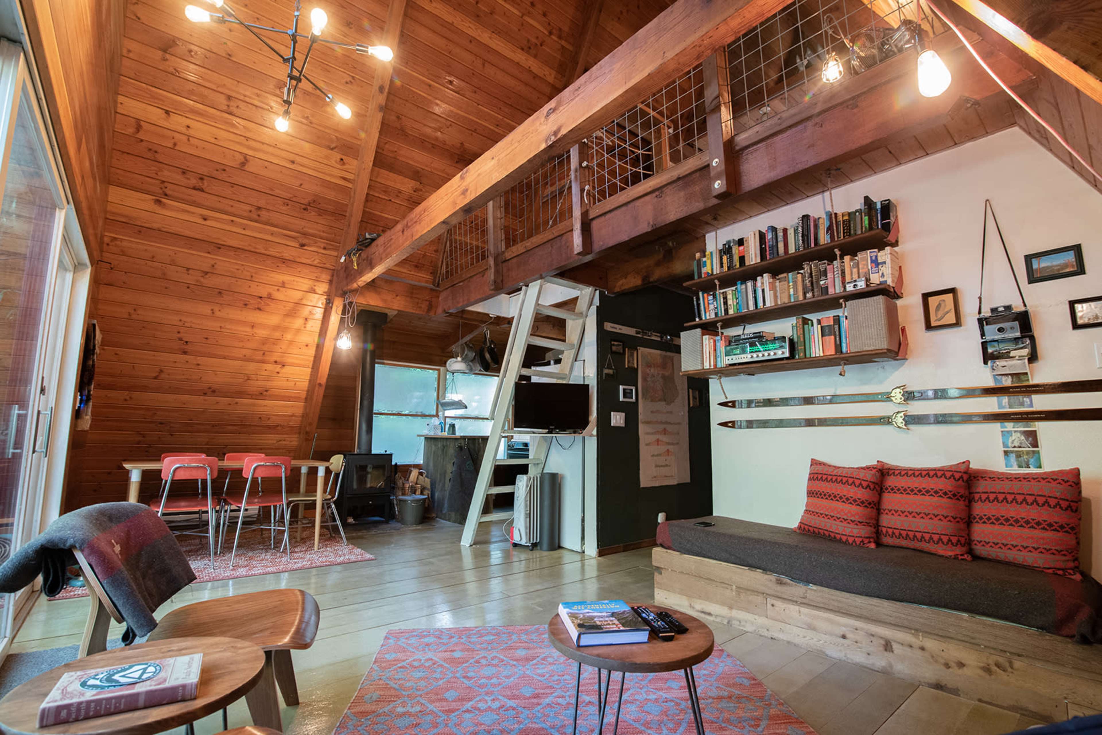A cozy, wooden interior of a cabin featuring a living area with a sofa, a dining table, and a lofted sleeping space.