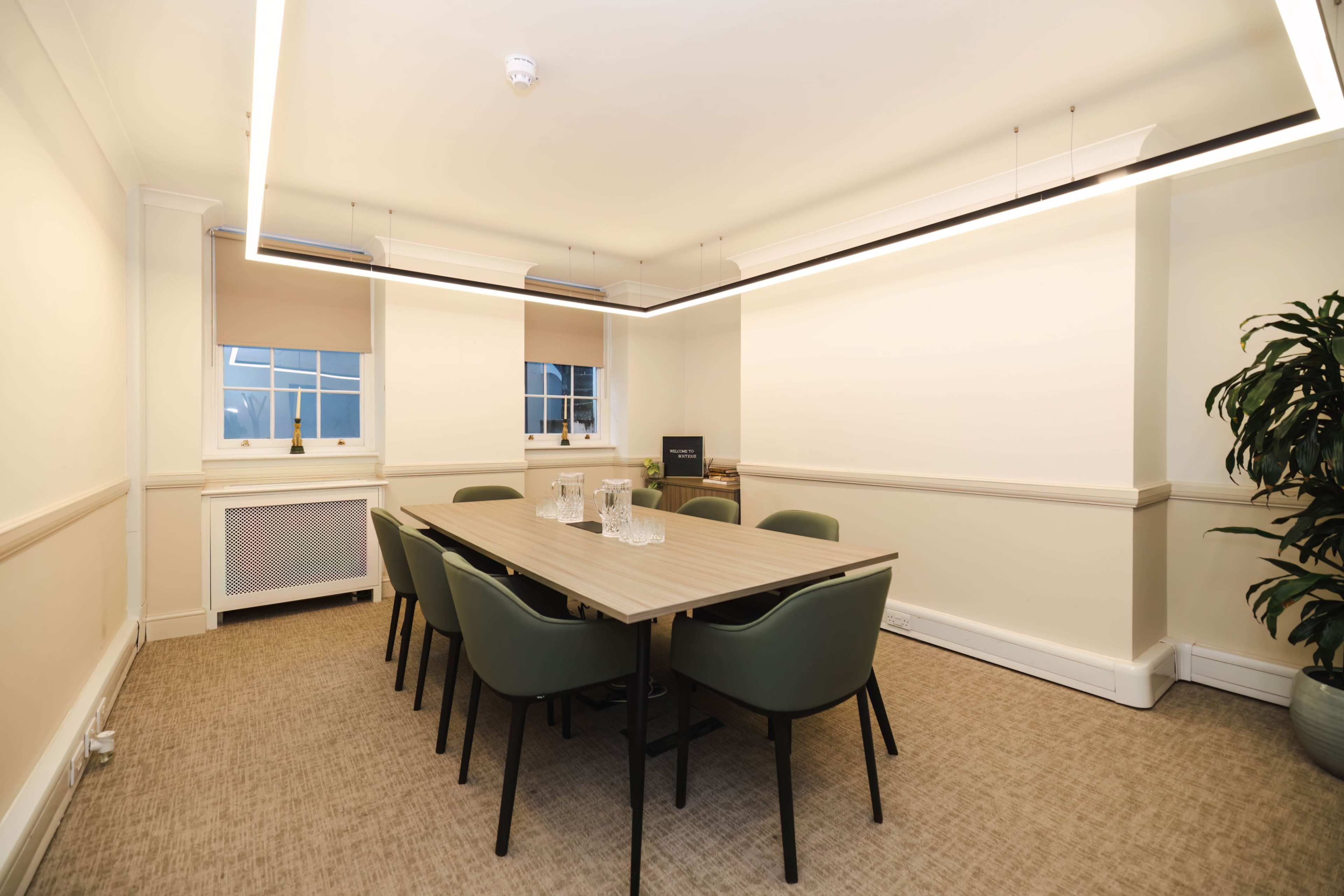 A well-lit meeting room with a rectangular table surrounded by six chairs, featuring a plant in one corner and a large window with blinds.