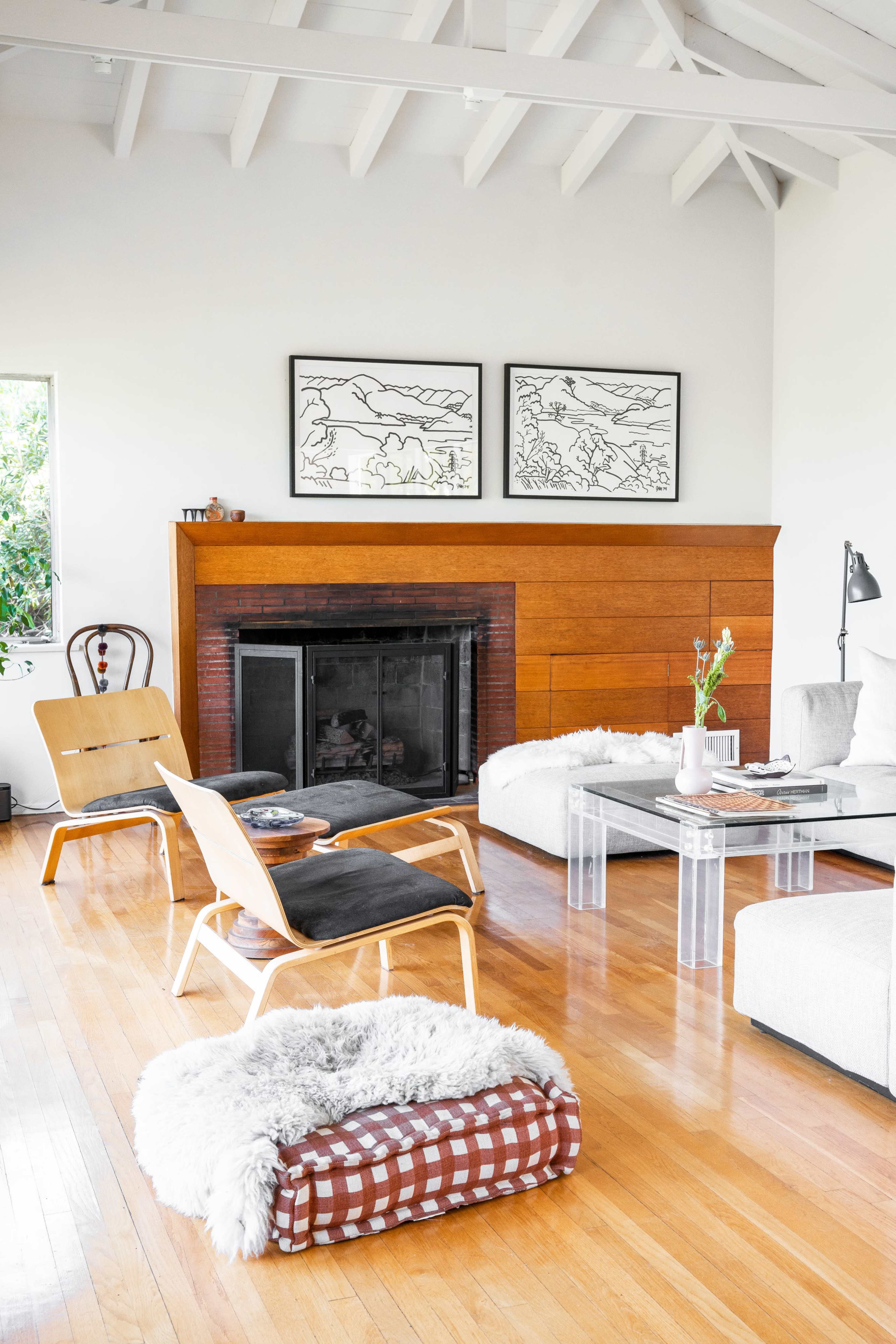 A living room features a fireplace surrounded by wood paneling, two distinct chairs, a glass coffee table, and framed artworks on the wall.