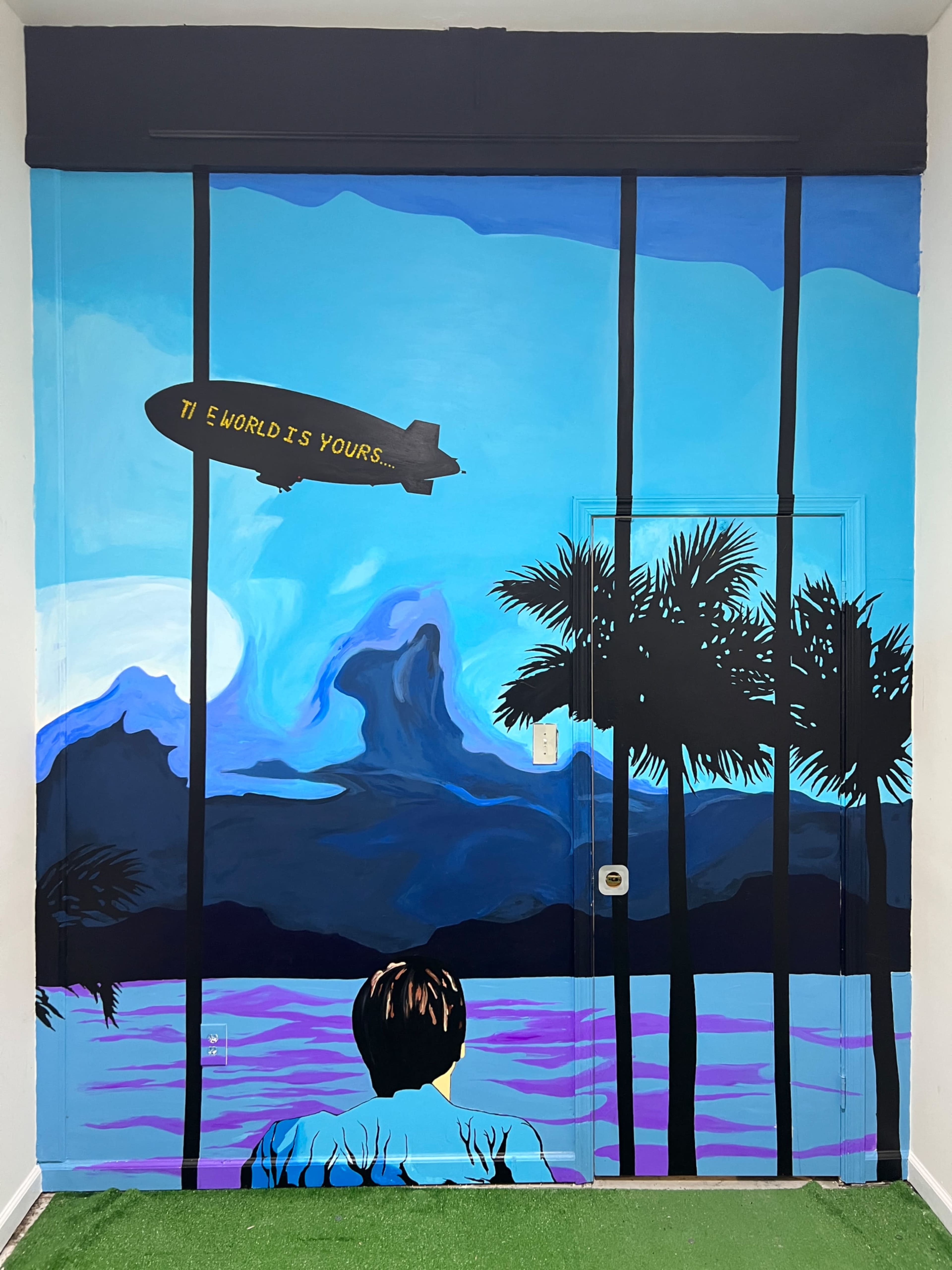 A mural of a boy looking out at a vibrant landscape with palm trees, mountains, and a blimp that reads "THE WORLD IS YOURS."