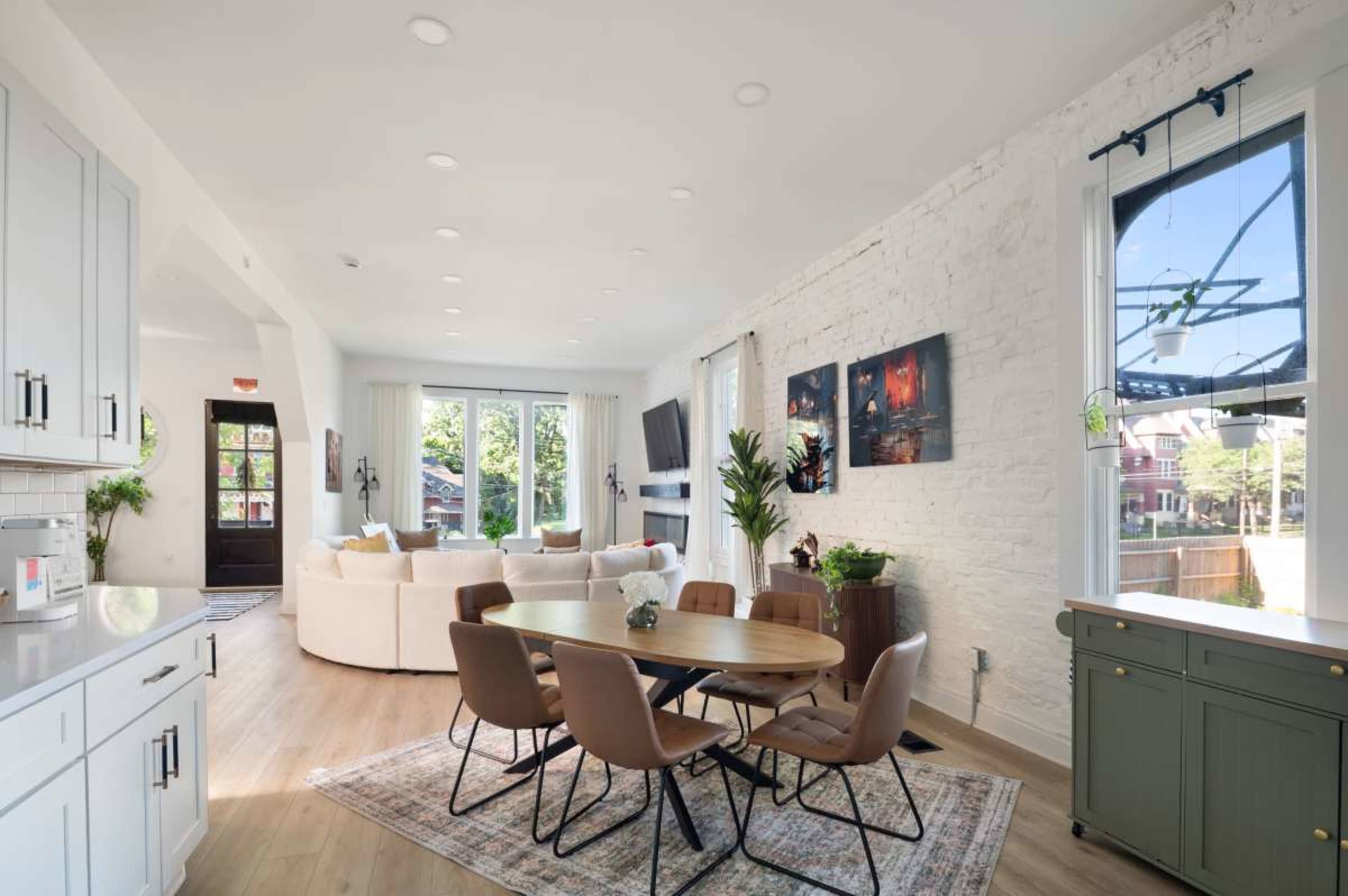 The image shows a modern open-concept living and dining area with a round wooden table surrounded by chairs, a white brick wall, and large windows allowing natural light.