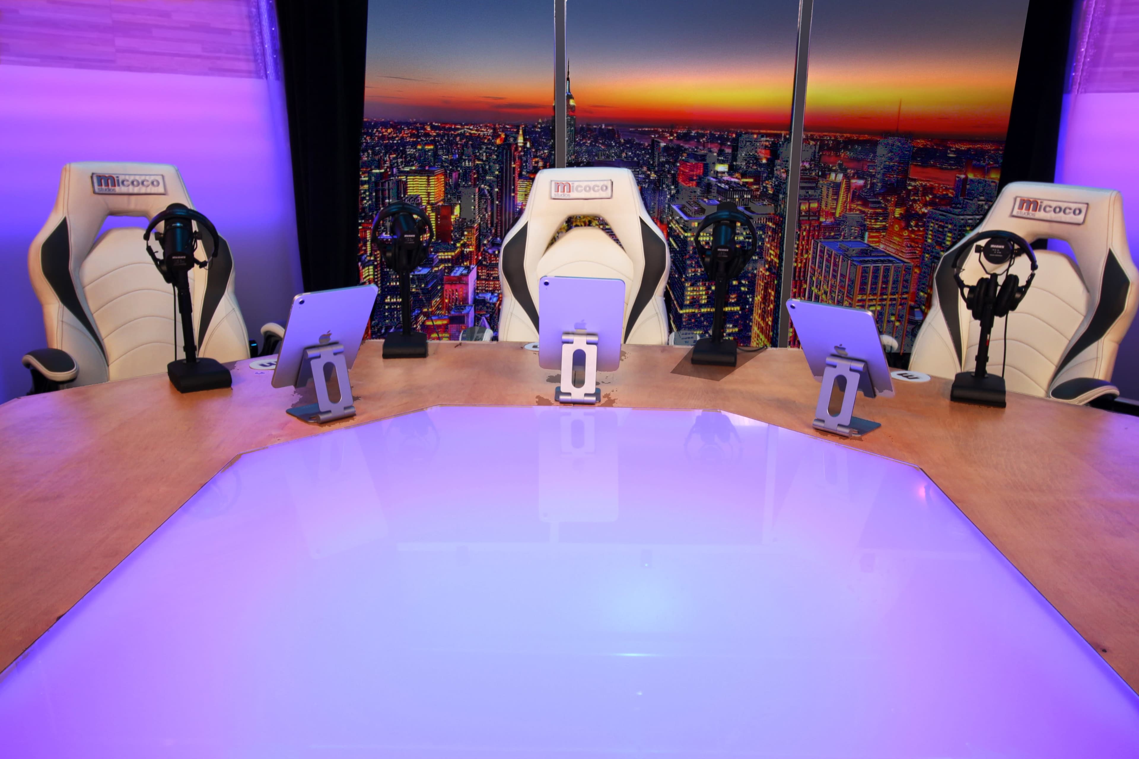 A modern conference room features a large table with four tablets and microphones, set against a vibrant city skyline view at sunset.