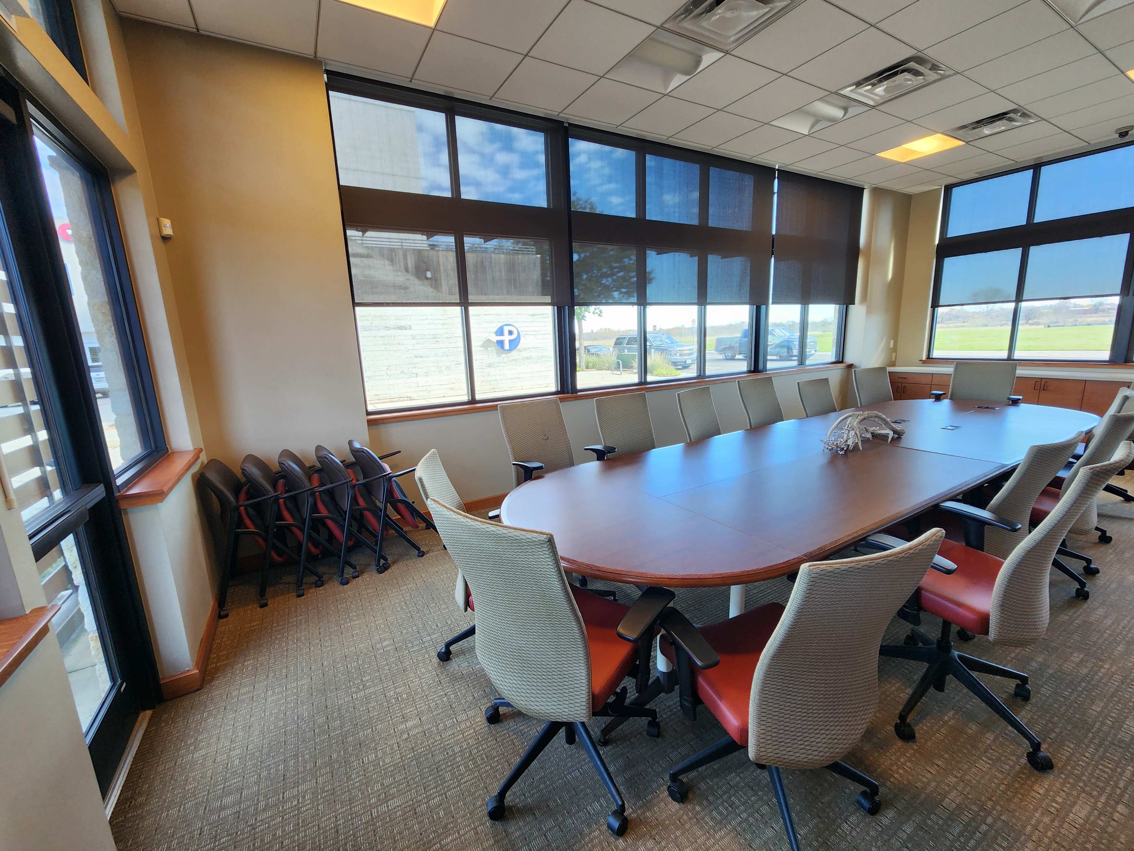 A conference room with a large oval table, rolling chairs, and windows providing natural light.