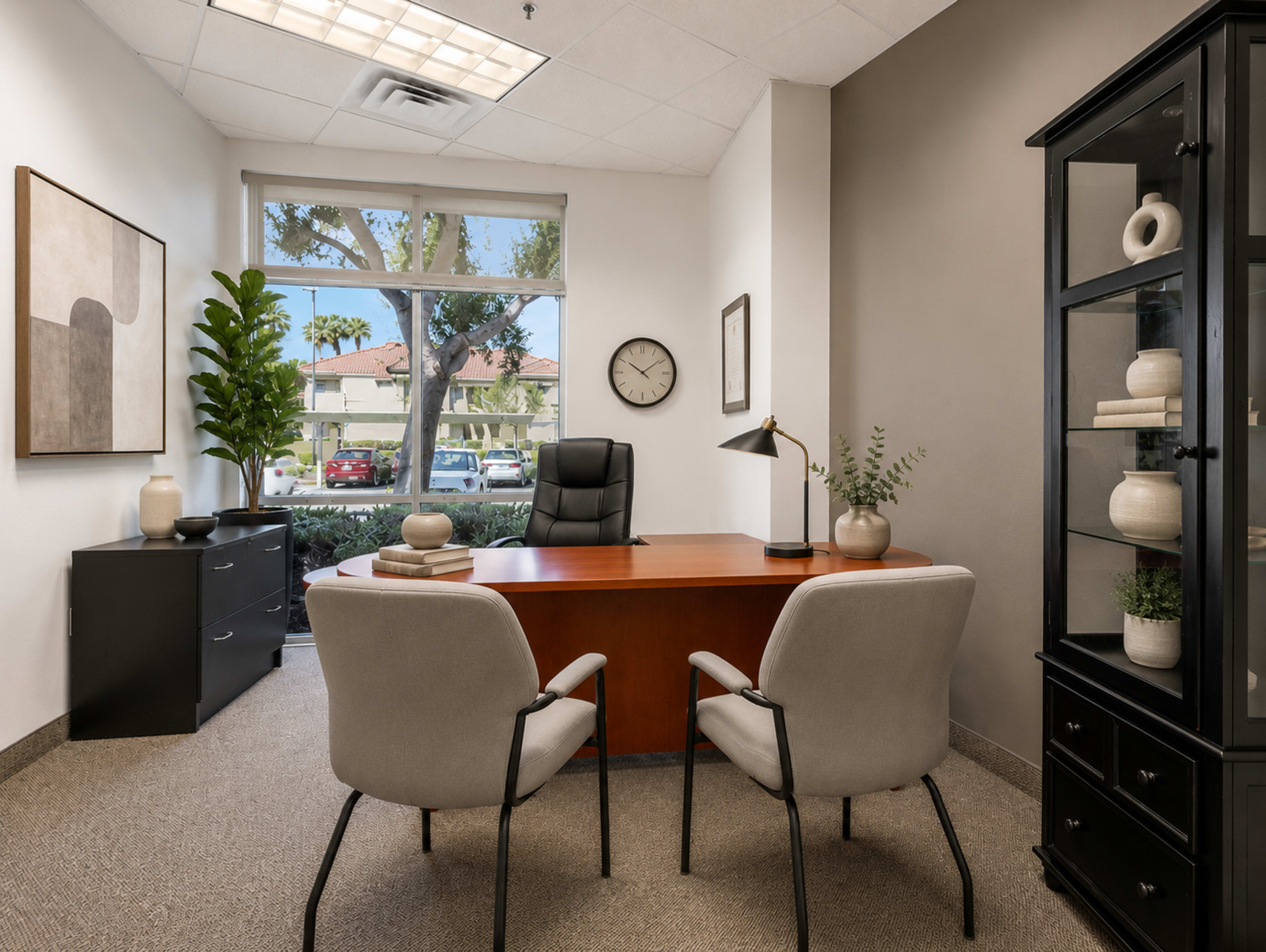 A modern office features a wooden desk, two chairs, and a large window with a view of a landscaped area outside.