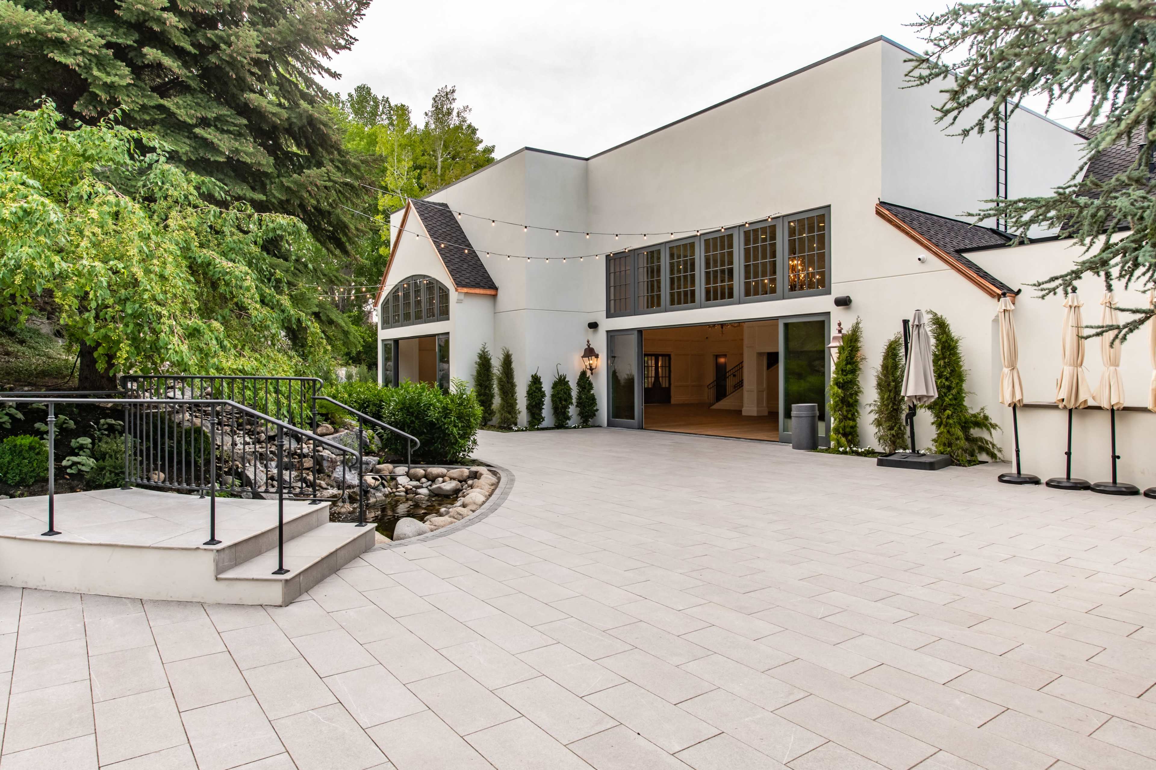 The image shows a modern building surrounded by greenery, featuring a paved outdoor area and a small stone water feature.