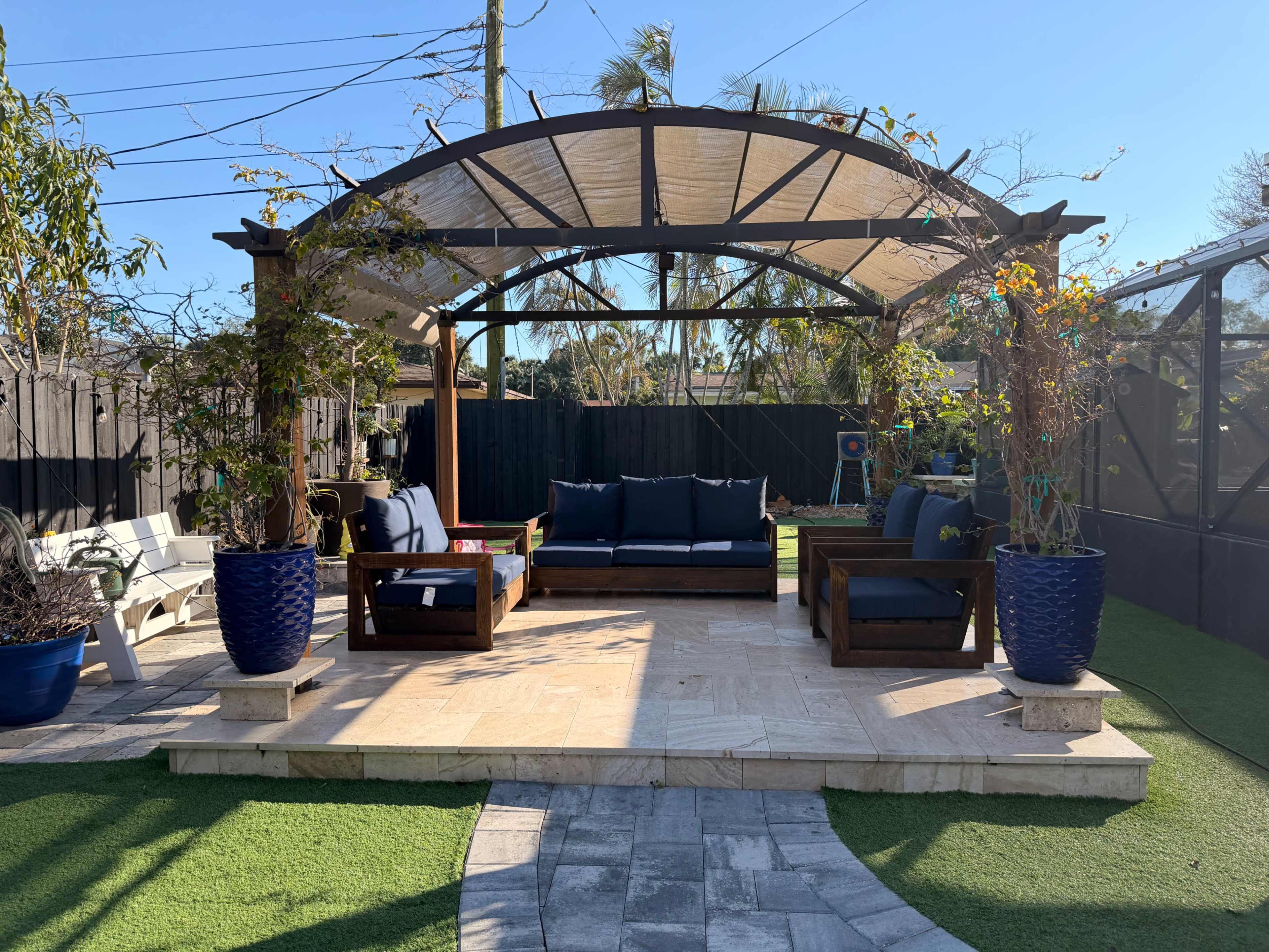 A well-designed outdoor seating area features a wooden pergola with seating and potted plants, surrounded by artificial grass and stone pathway.