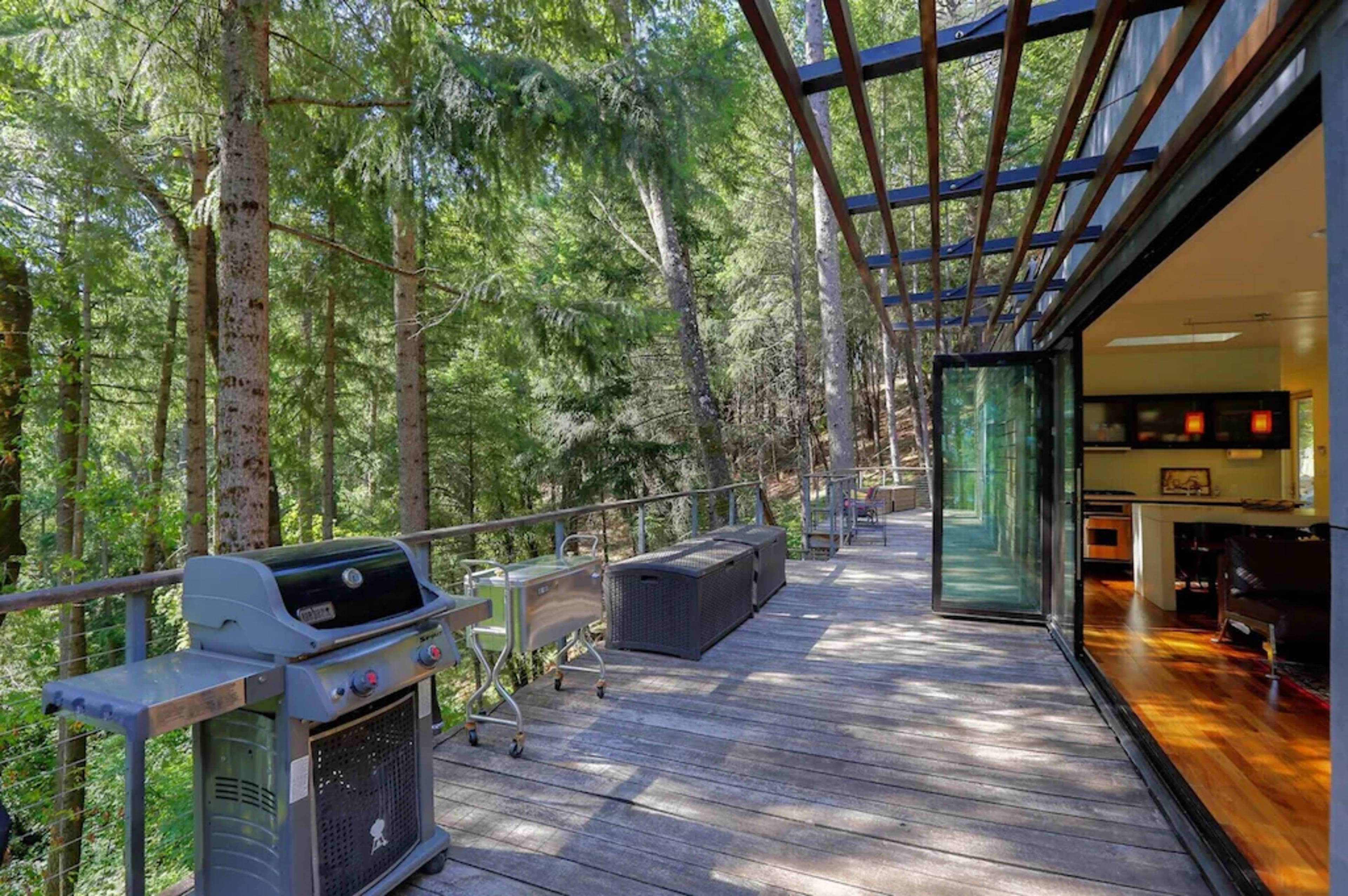 A wooden deck with a barbecue grill overlooks a wooded area, featuring lounge furniture and large glass doors leading into a modern interior.