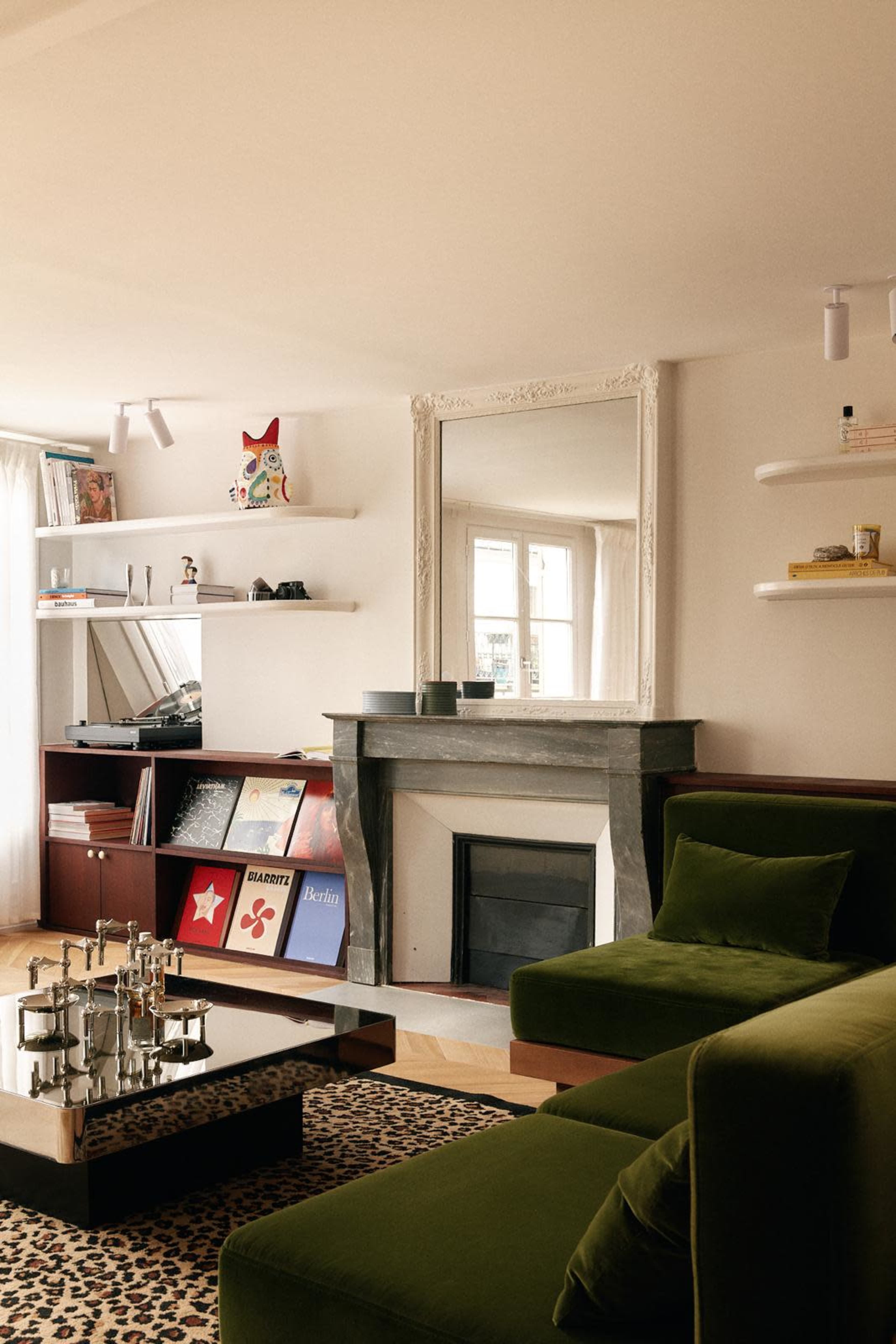 The image shows a modern living room featuring a green velvet sofa, a coffee table, and a fireplace with a marble surround, alongside shelves filled with records and decorative items.