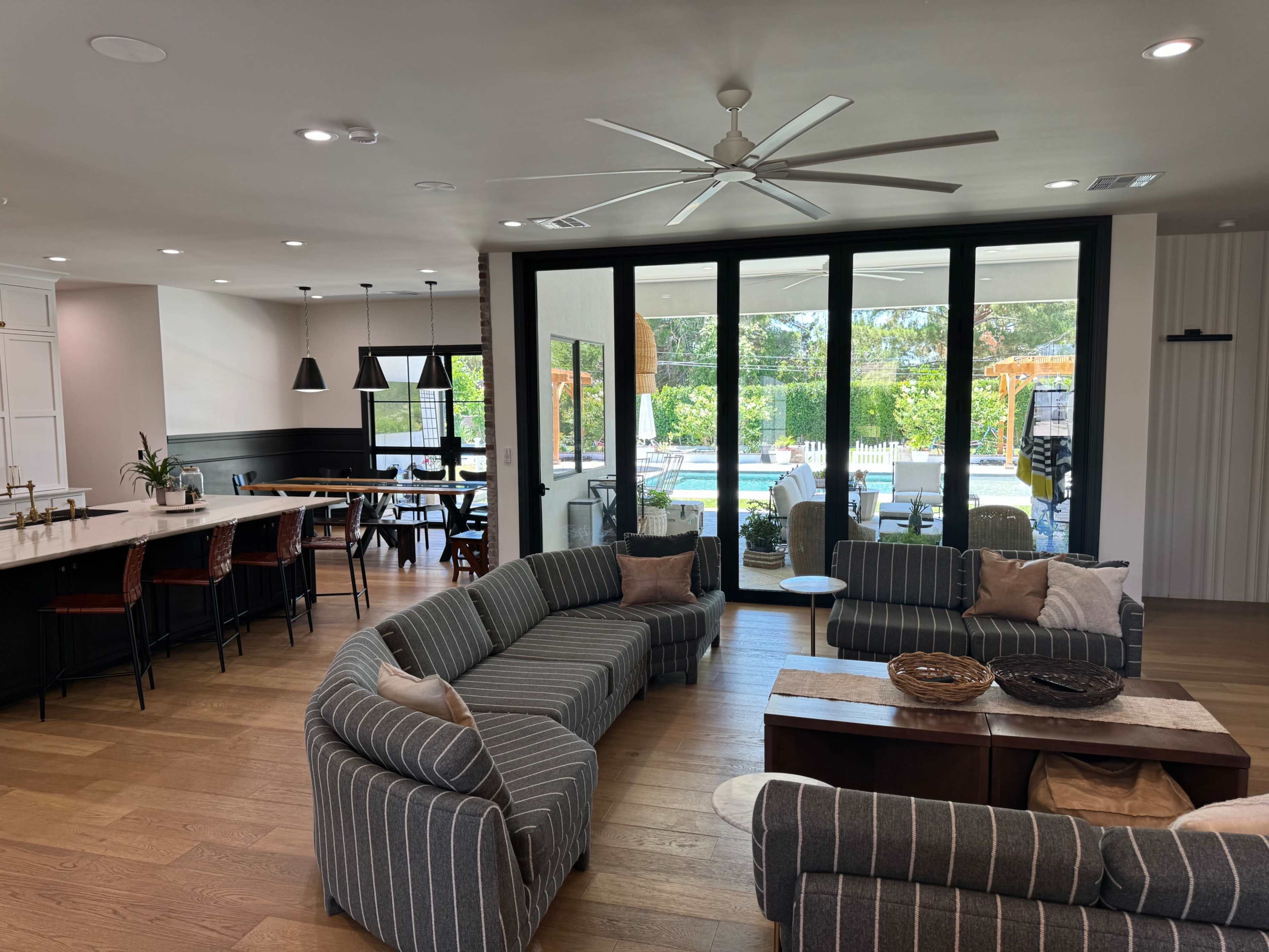 A spacious, modern living area with striped sofas arranged around a wooden coffee table, leading to a large glass door that opens to a poolside patio.