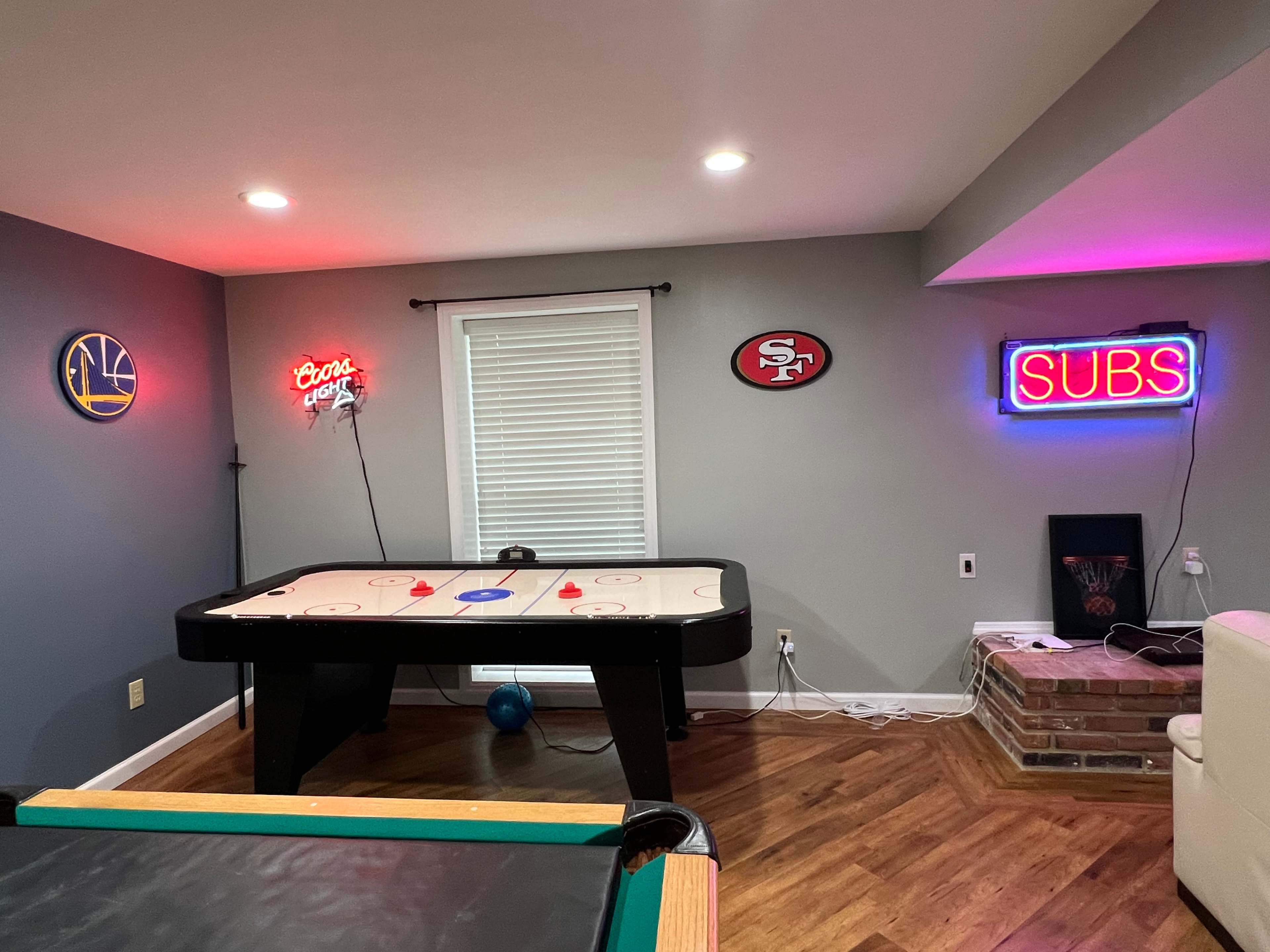 A game room featuring an air hockey table, a pool table, and neon signs indicating "SUBS" and a logo, with wood flooring and a window covered by blinds.