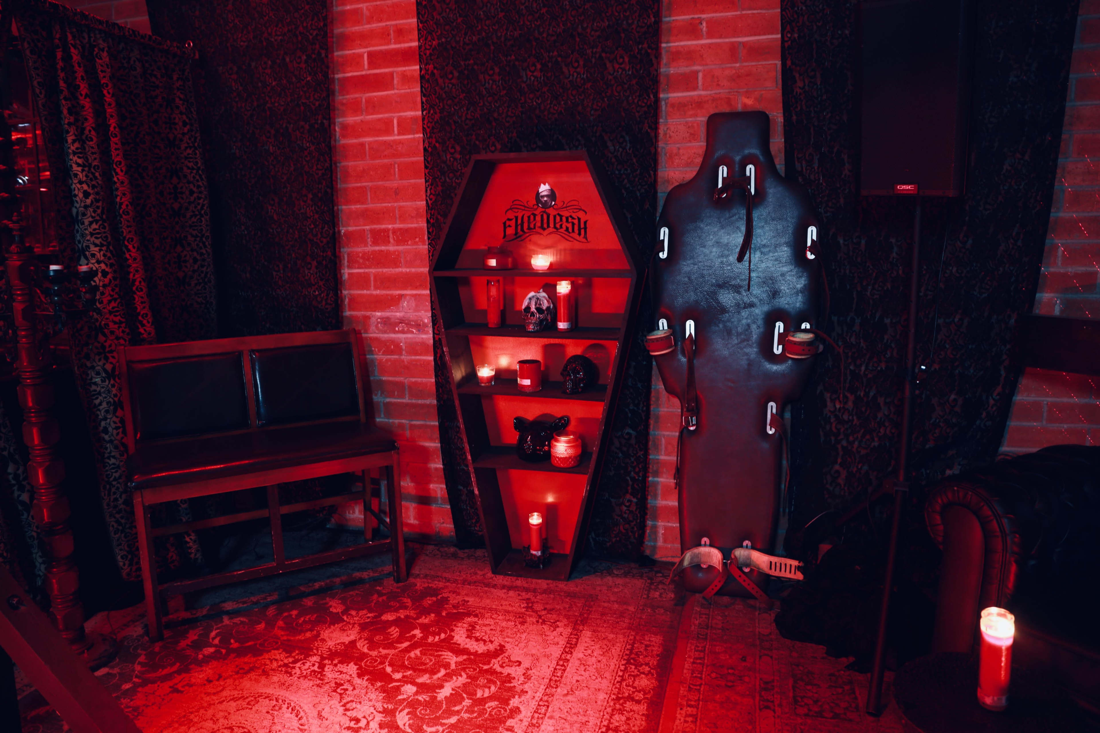 The image depicts a dimly lit room featuring a wooden coffin-shaped shelf with various objects, a black padded table, and candles, all set against a backdrop of red lighting and dark drapes.