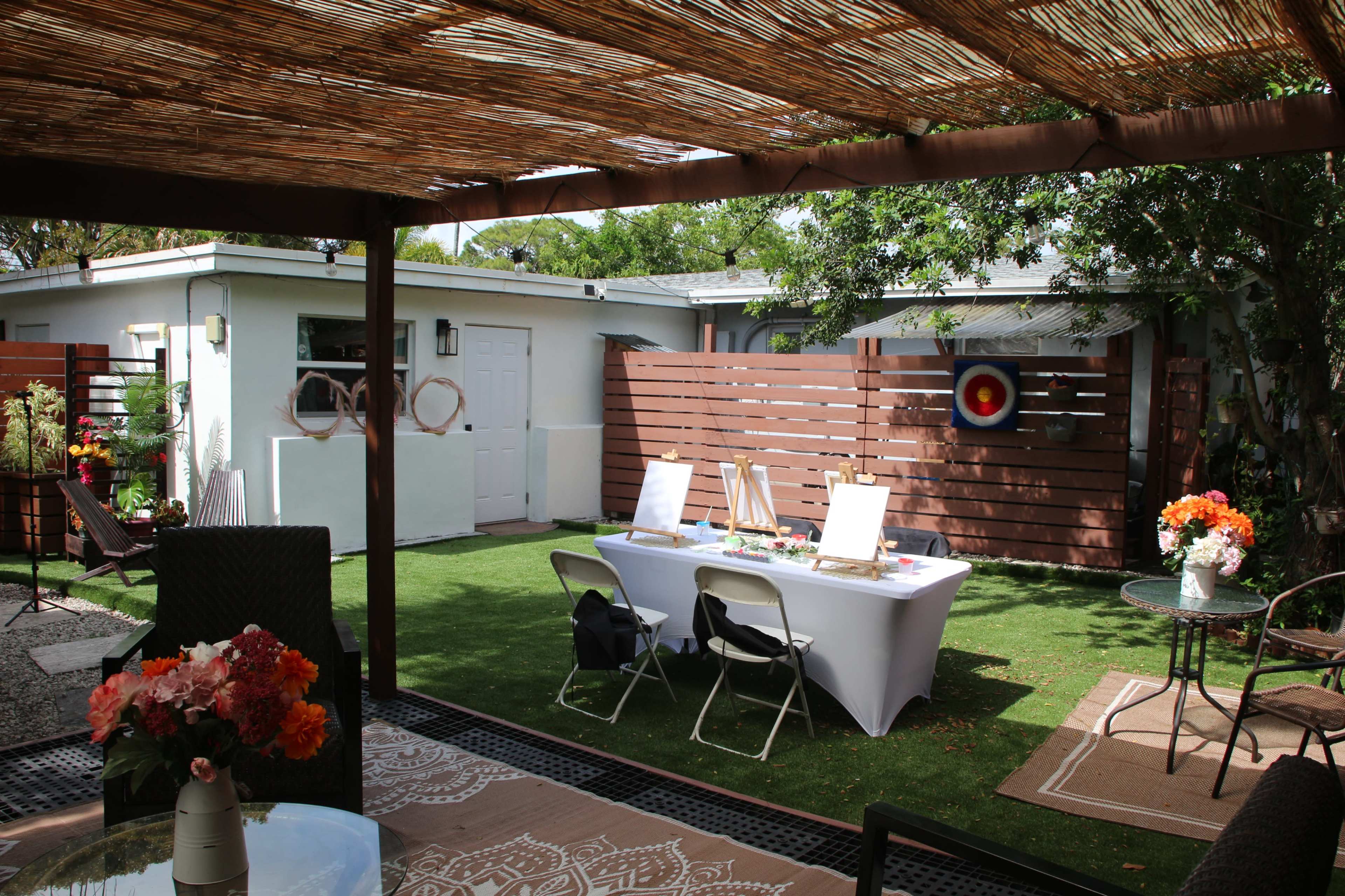 The image shows a backyard with a patio area featuring a table set for painting, surrounded by greenery and wooden fencing.