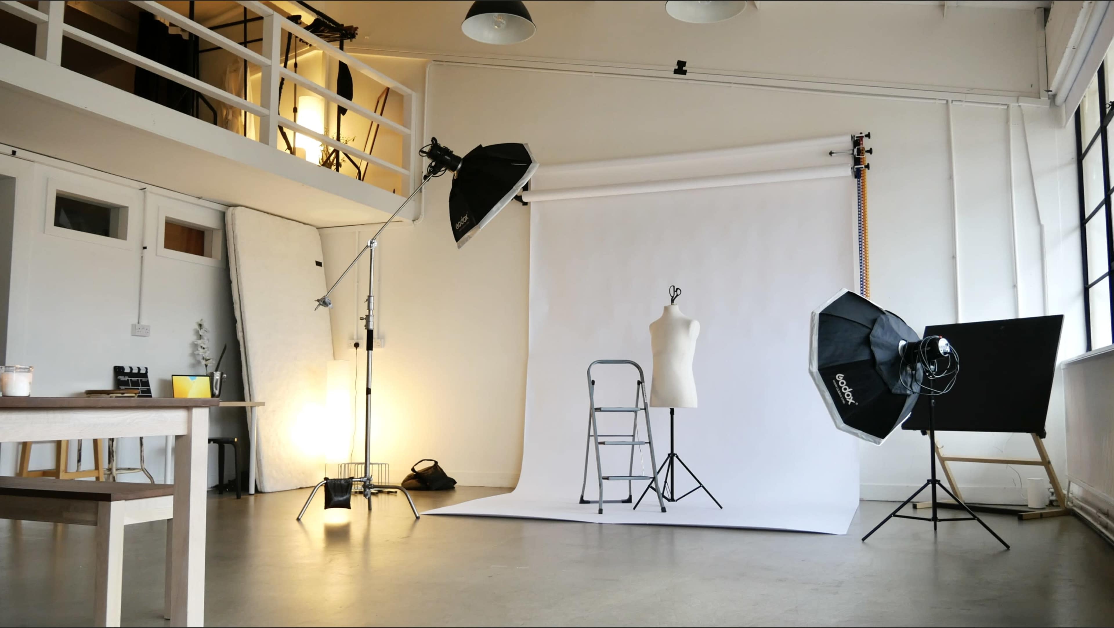 The image shows a photography studio with a white backdrop, a mannequin, lighting equipment, and a ladder set up for a photoshoot.