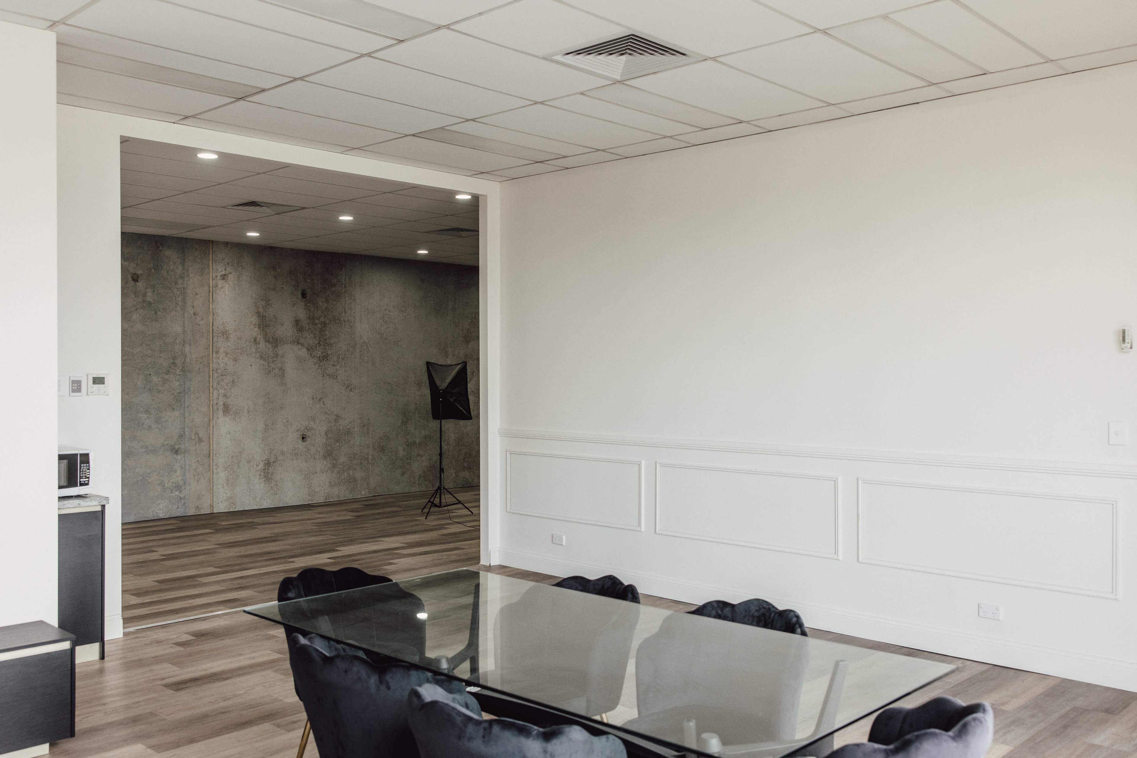 A modern office space features a glass dining table surrounded by black chairs, with an adjacent room showcasing a concrete wall and studio lighting setup.