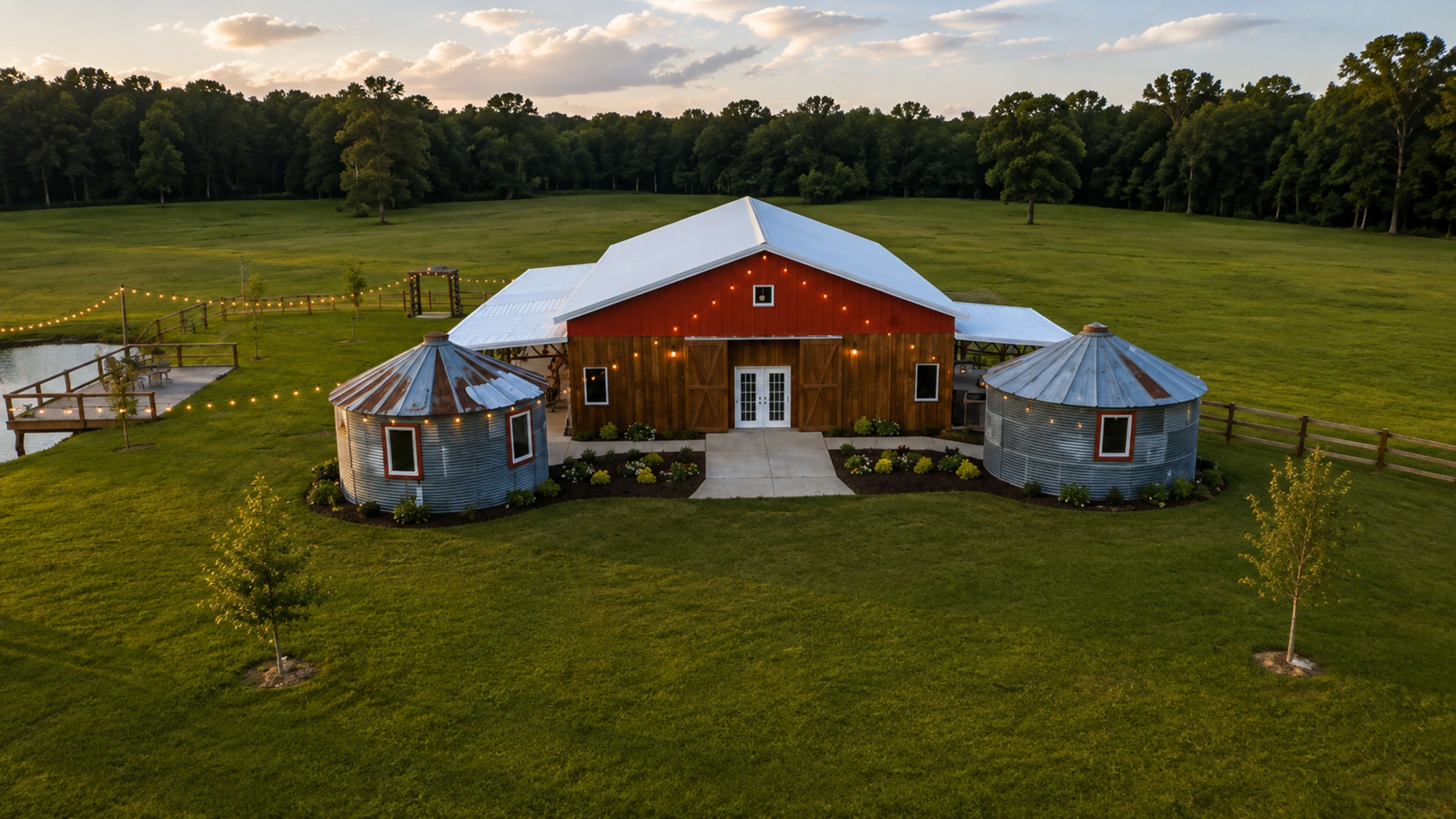 The image shows a barn-style building with a red wooden facade, surrounded by green grass and two silver silos on either side.