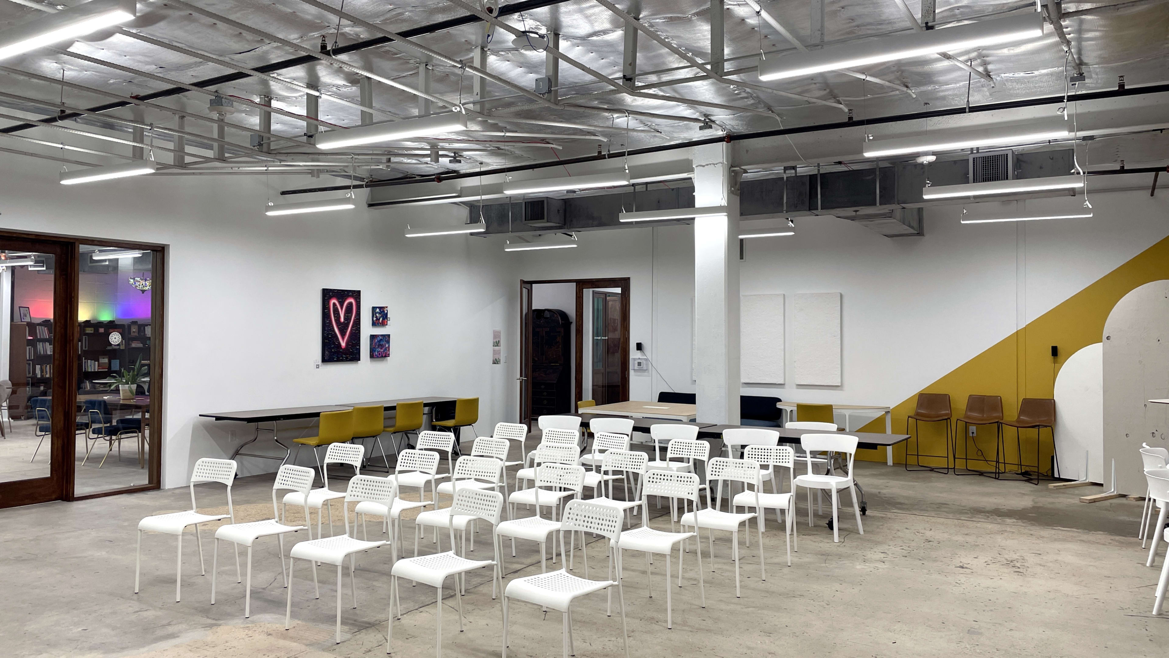 The image shows a minimally furnished room with white chairs arranged in rows, a long table, and colorful wall art.