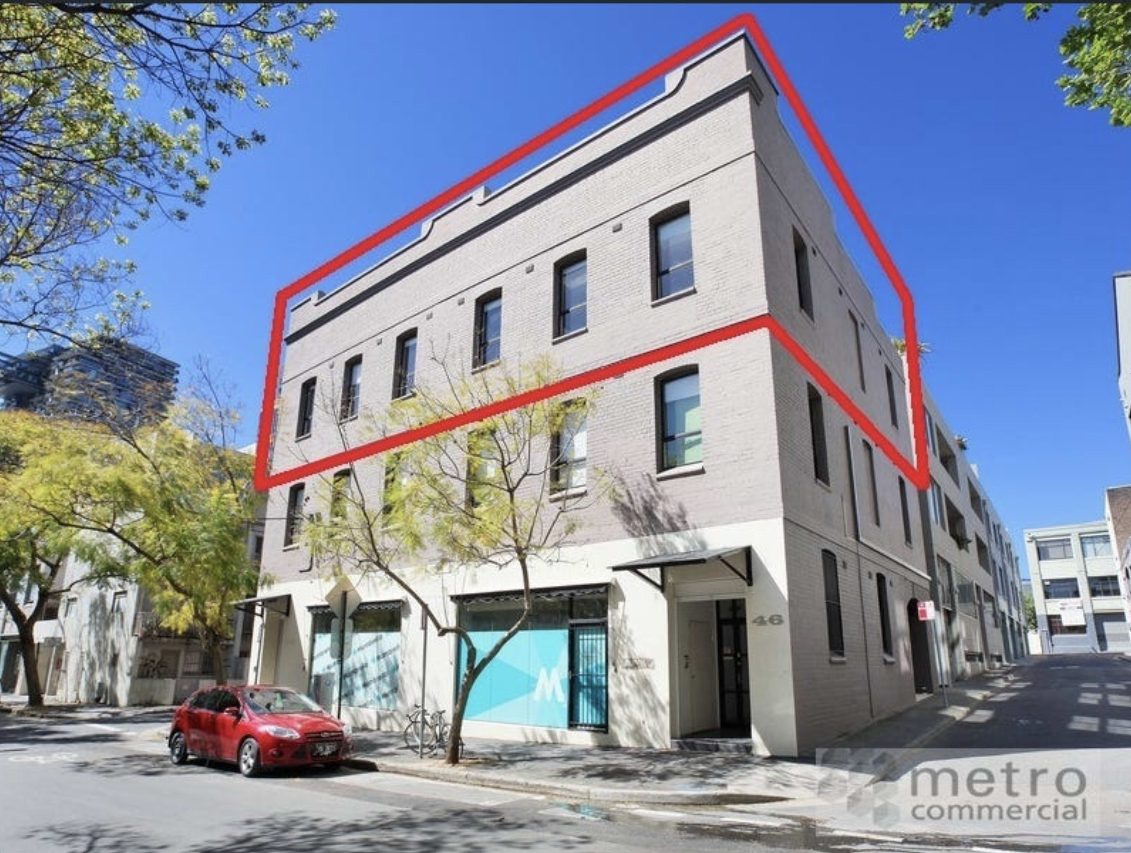 A three-story commercial building with large windows is located on a street corner, surrounded by trees and a parking area.
