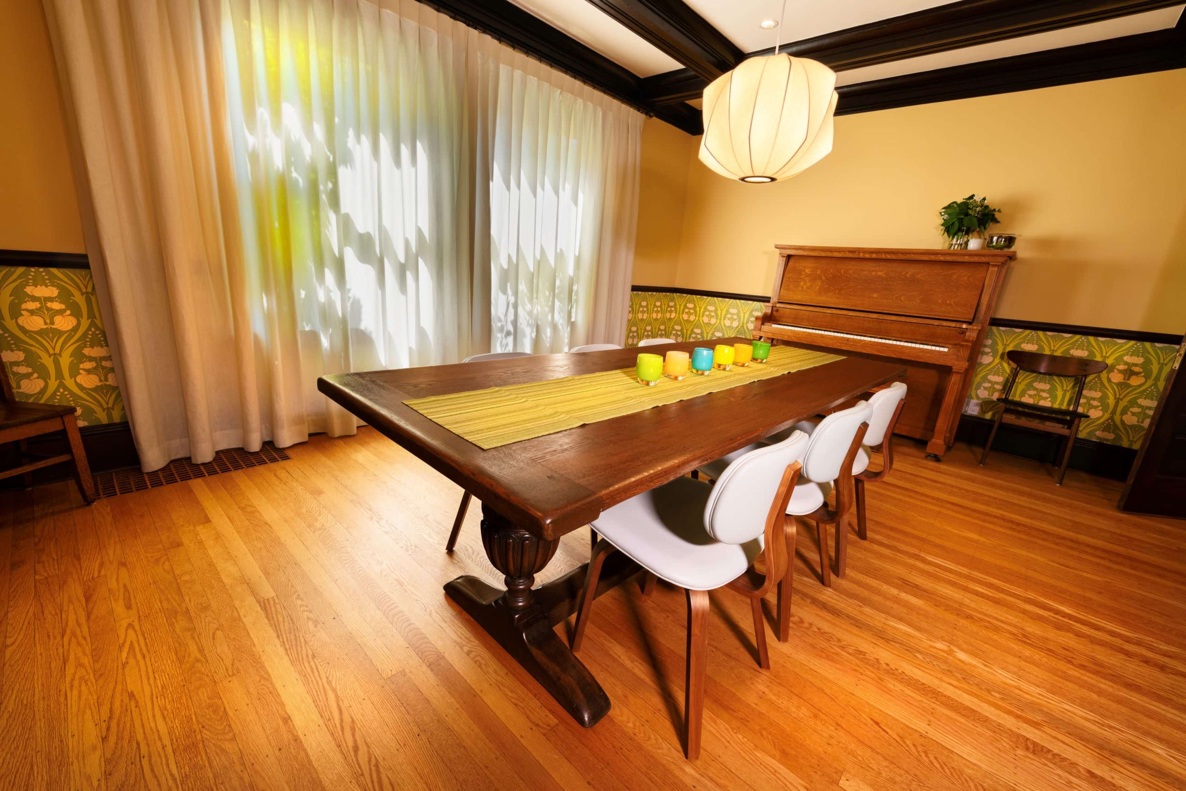 A spacious dining room features a large wooden table set with colorful cups, surrounded by modern chairs, with a piano and patterned wall decor in the background.