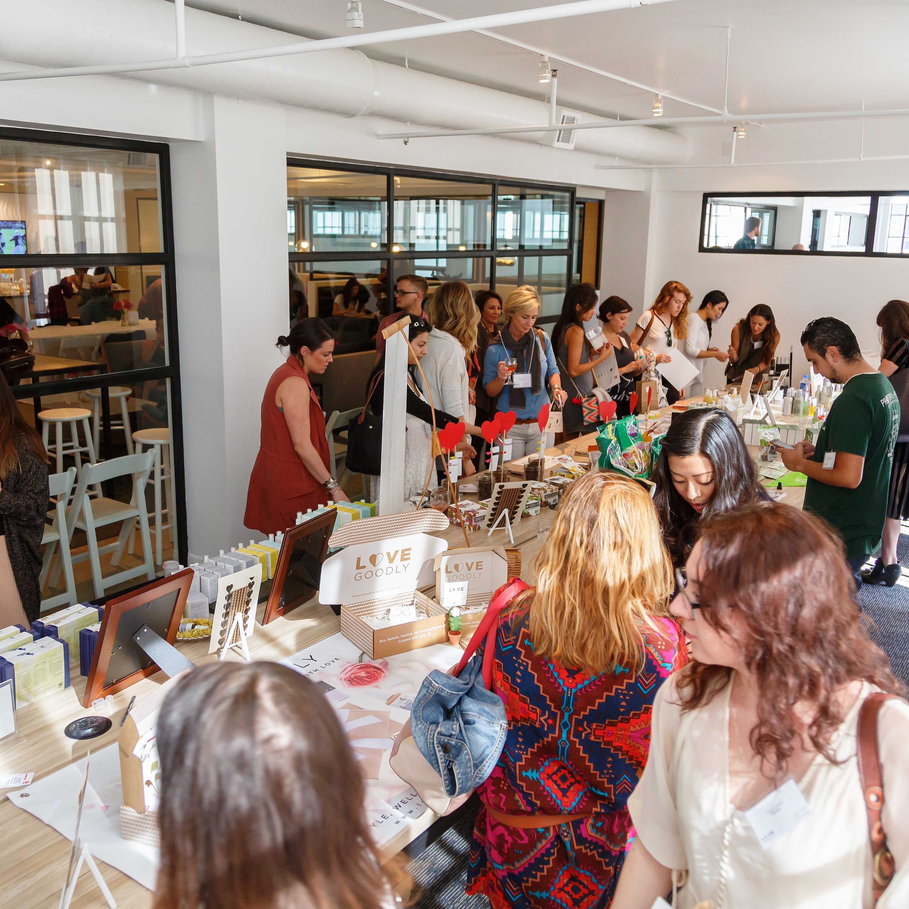 A diverse group of people gathers around tables displaying various products in a bright, open room.