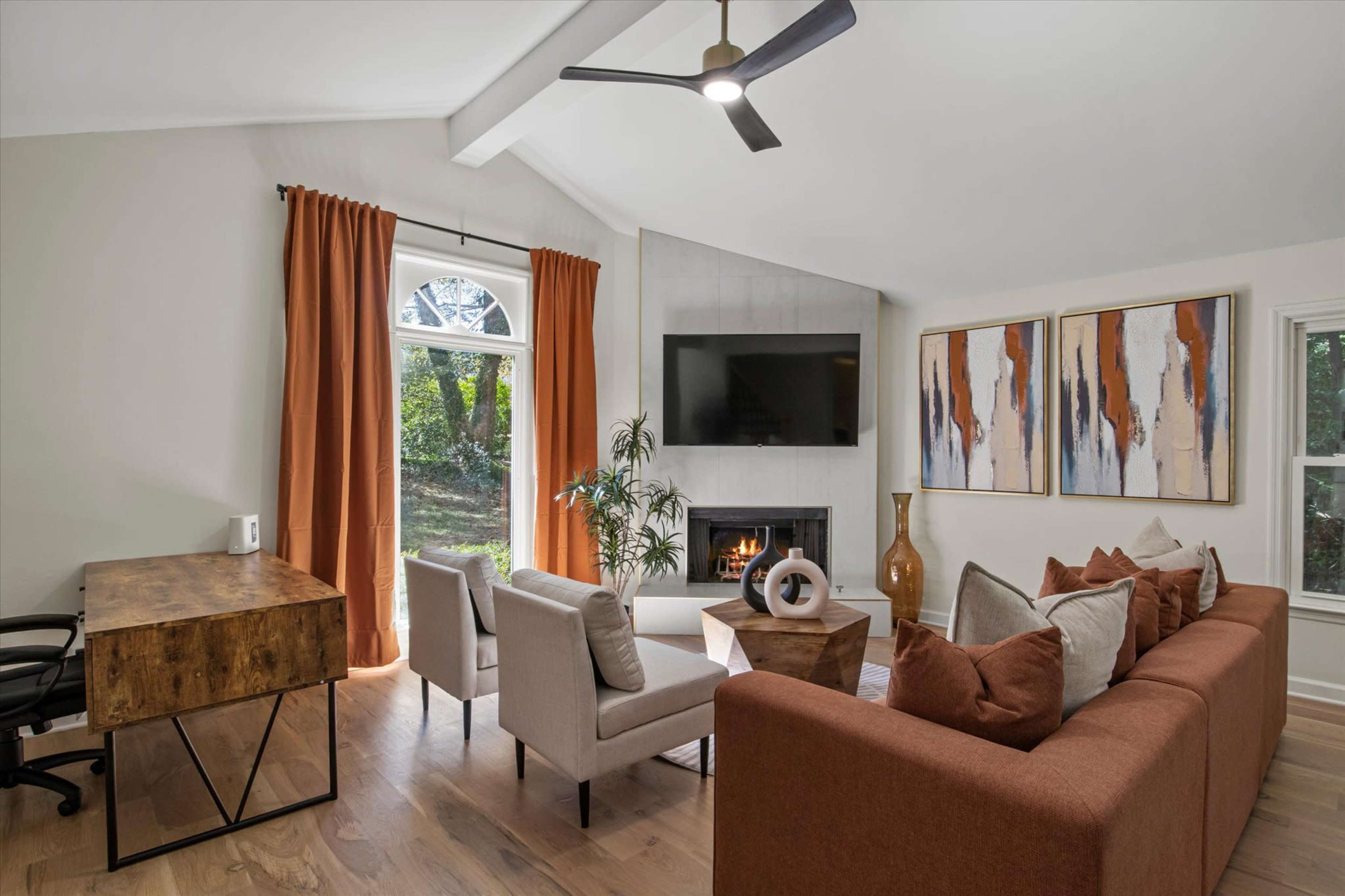 The living room features a modern layout with a fireplace, large windows, a comfortable sofa, and a desk.