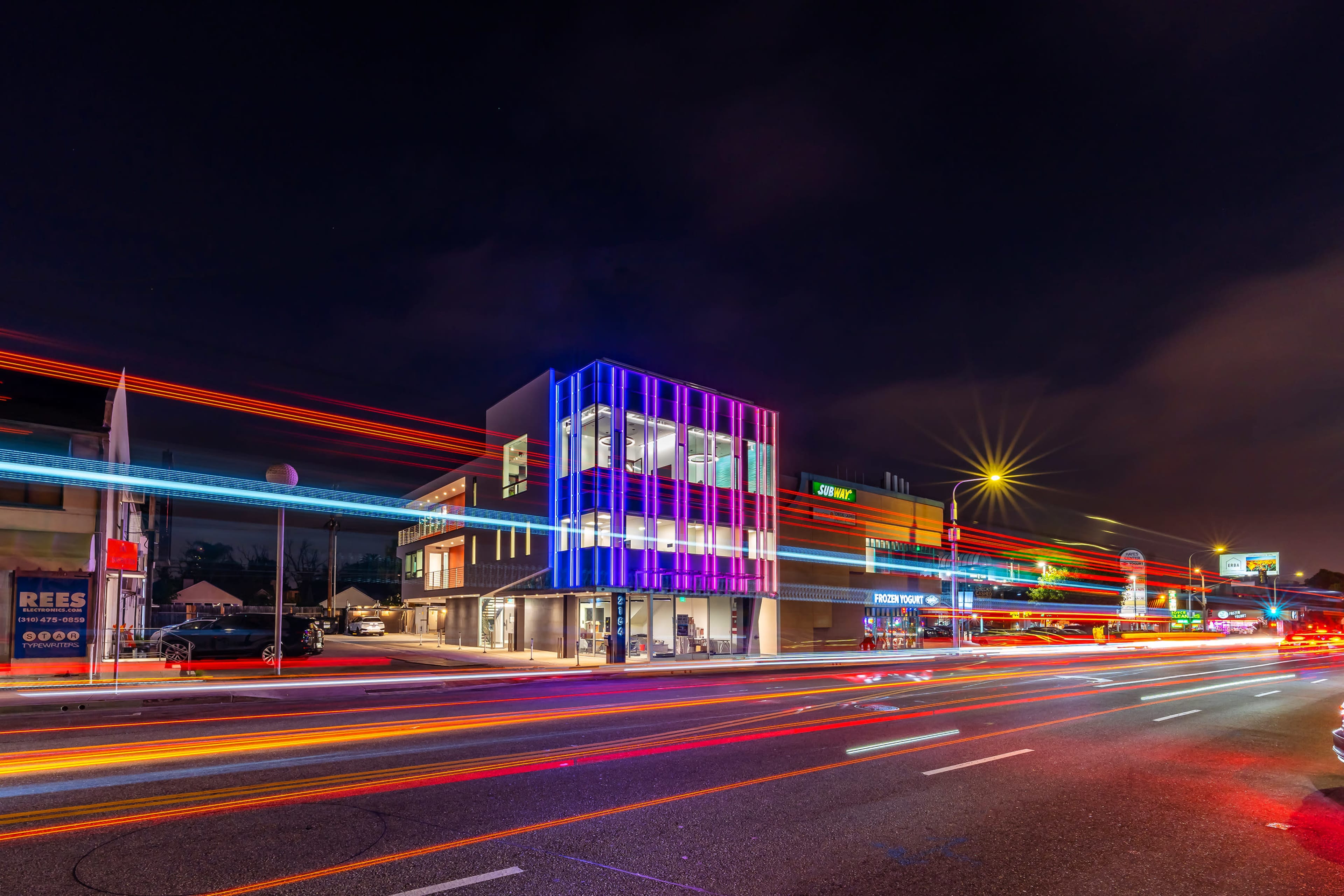 A modern building with colorful light accents stands beside a busy road at night, with light trails from passing vehicles creating a dynamic effect.