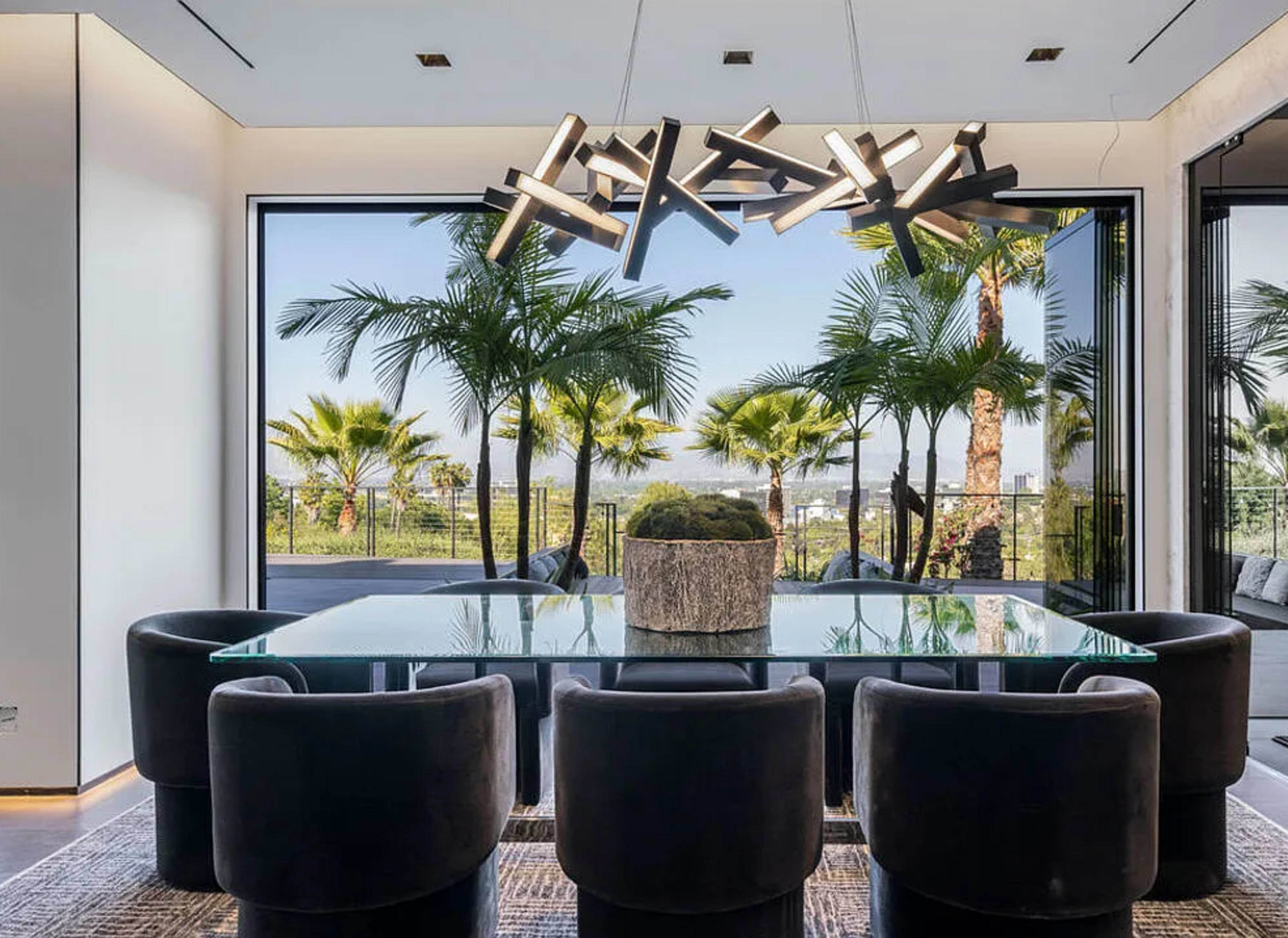 A modern dining room features a large glass table surrounded by six black upholstered chairs, with a striking chandelier overhead and large windows that frame a view of palm trees outside.