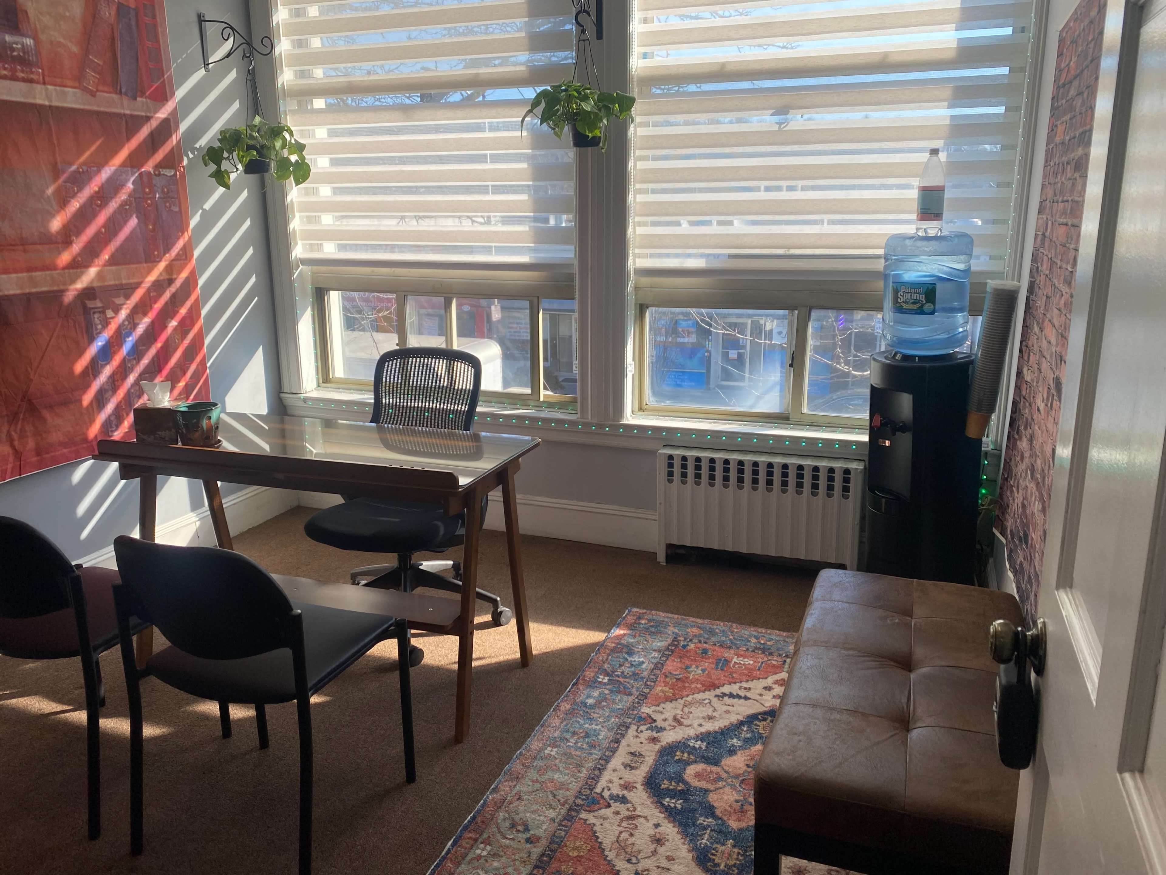 A small office space features a wooden table, black chairs, a water cooler, and large windows with blinds, illuminated by natural light.