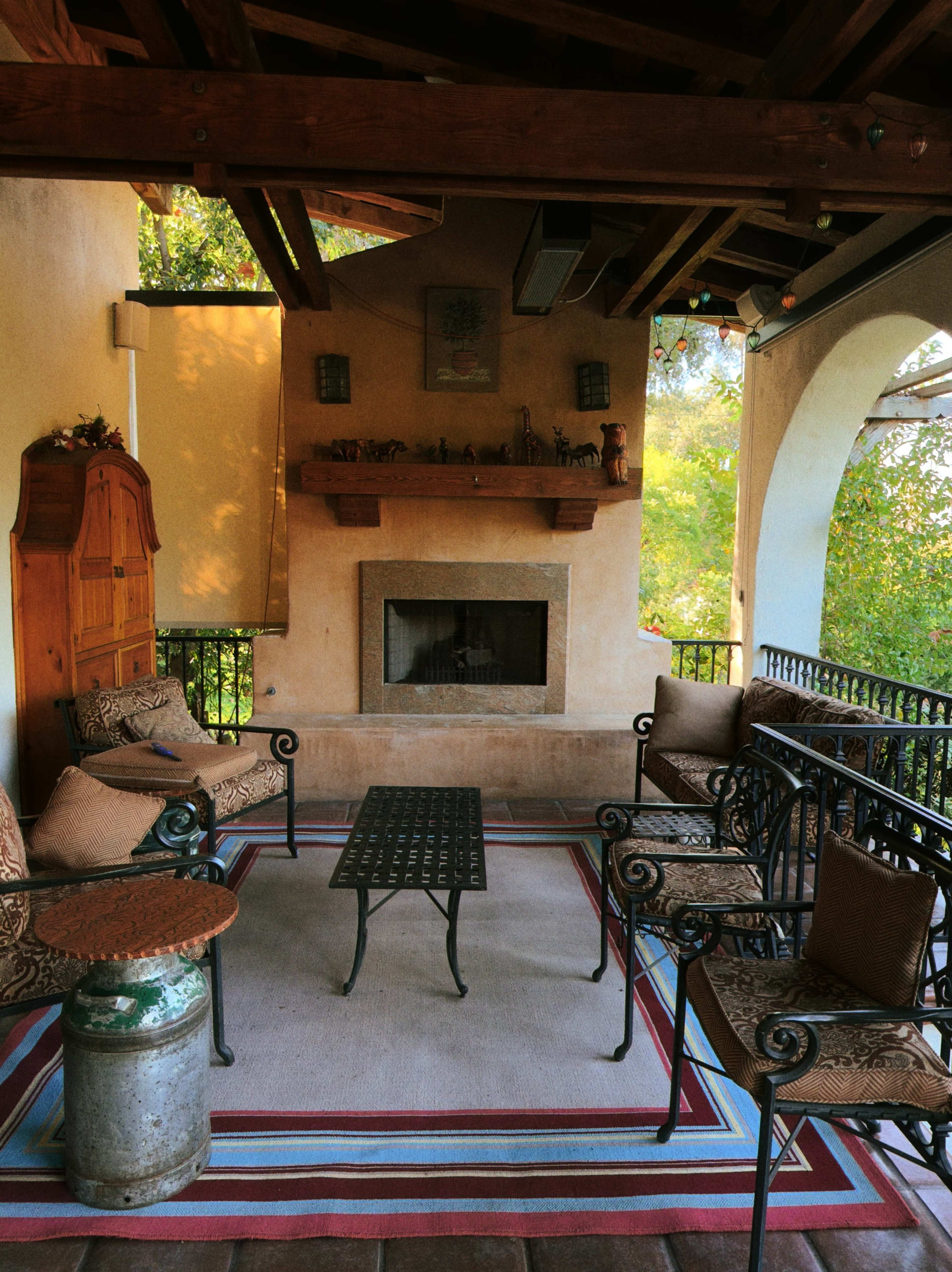 The image shows a cozy covered patio with a fireplace, seating area, and a rug, surrounded by greenery.