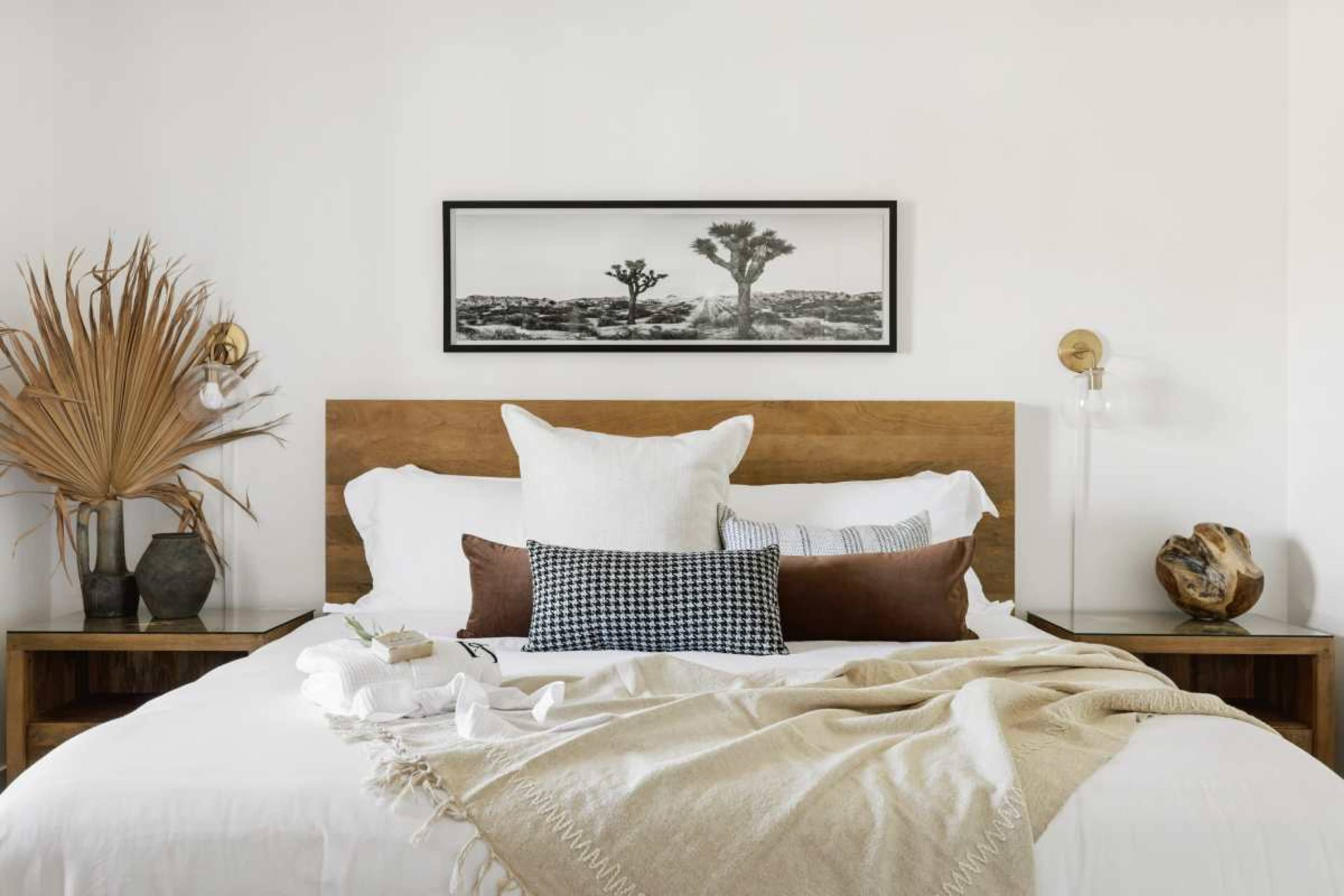 The image shows a neatly arranged bed with white linens, multiple decorative pillows, a wooden headboard, bedside tables, and a framed black and white photograph on the wall above.