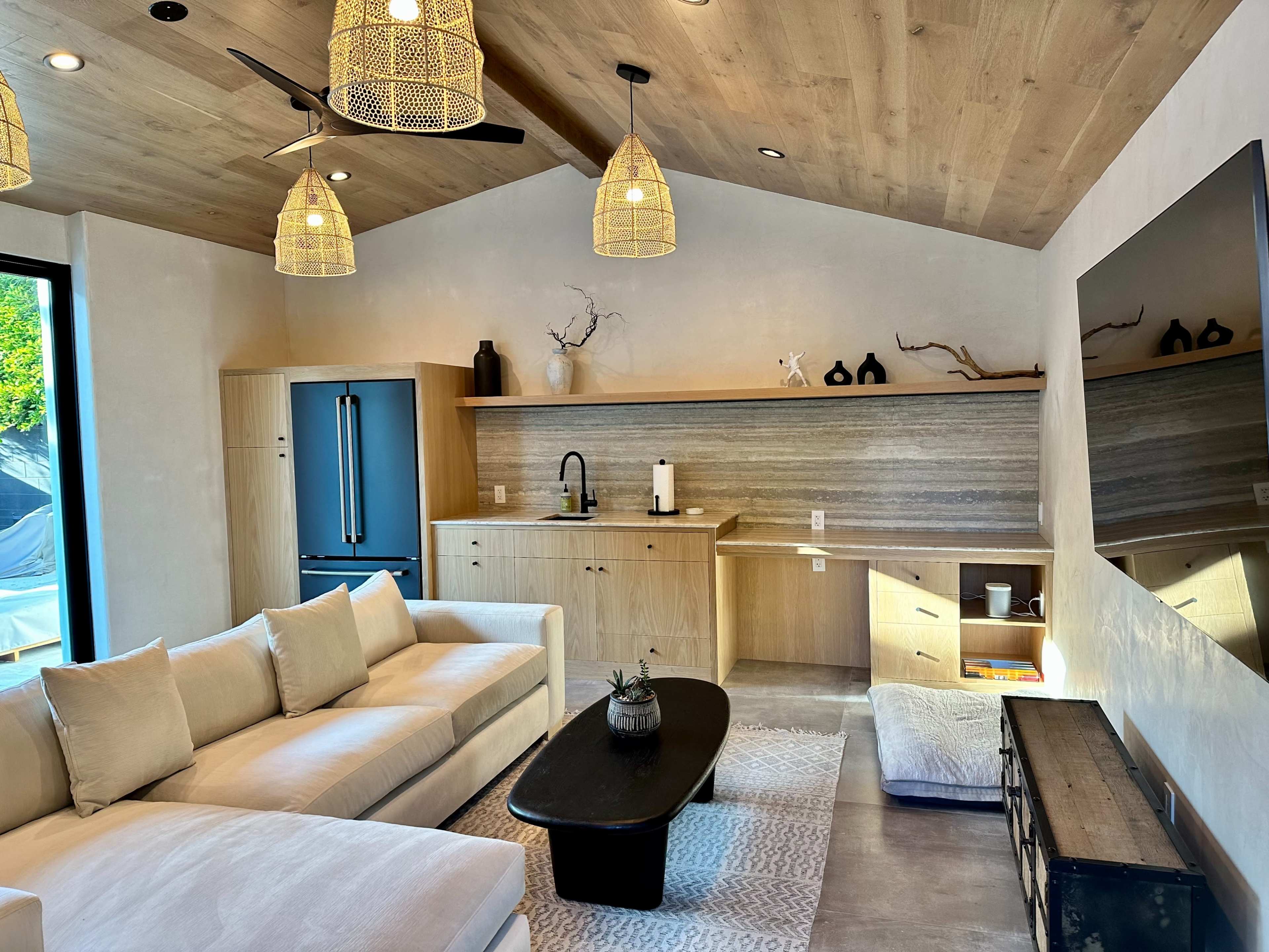 A modern living room with a sectional sofa, a black coffee table, a kitchen area with a refrigerator, and natural wood accents throughout.