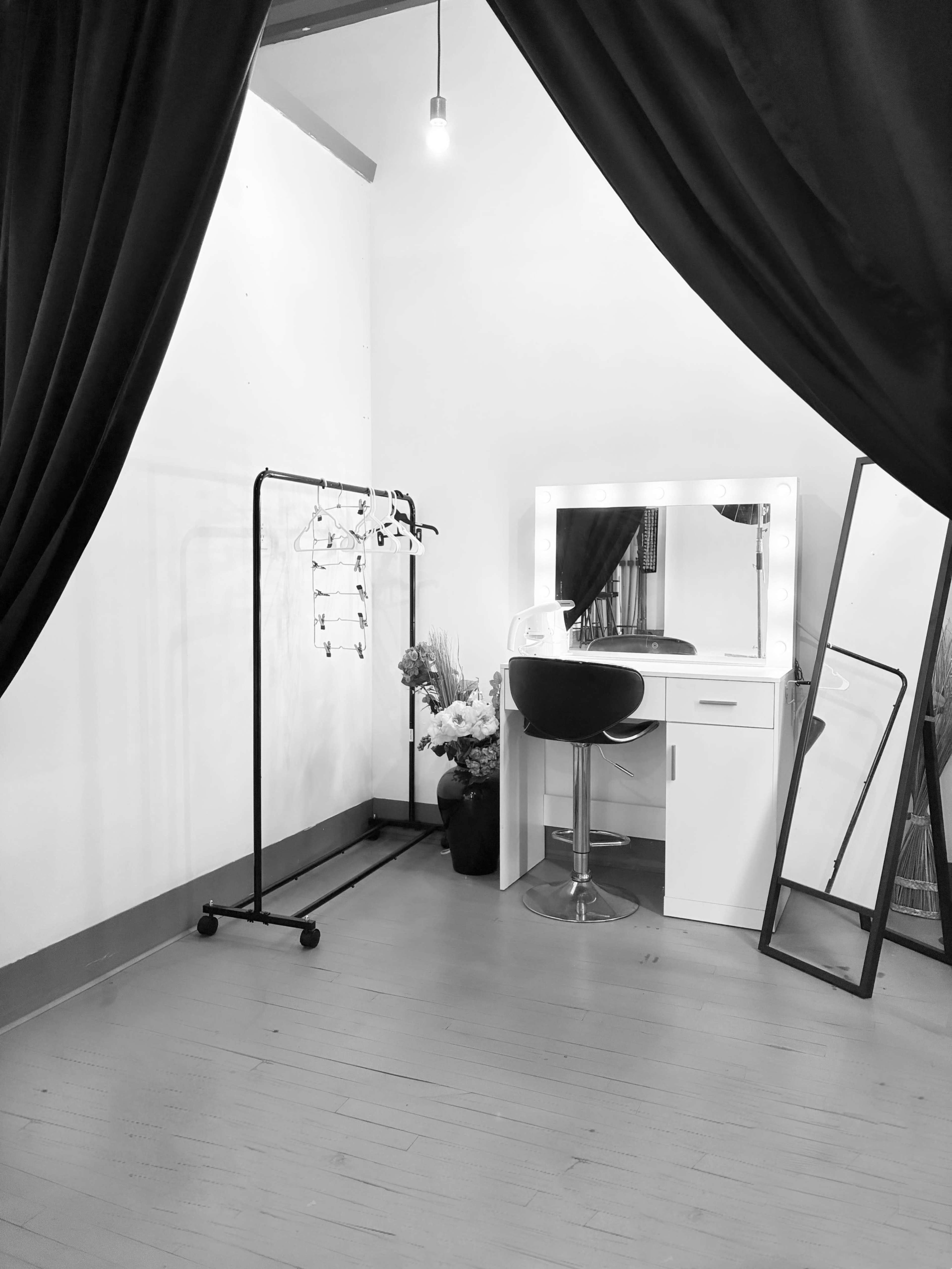 The image shows a vanity setup with a mirror, stool, and a rack for clothing, all within a space partially draped by black curtains.