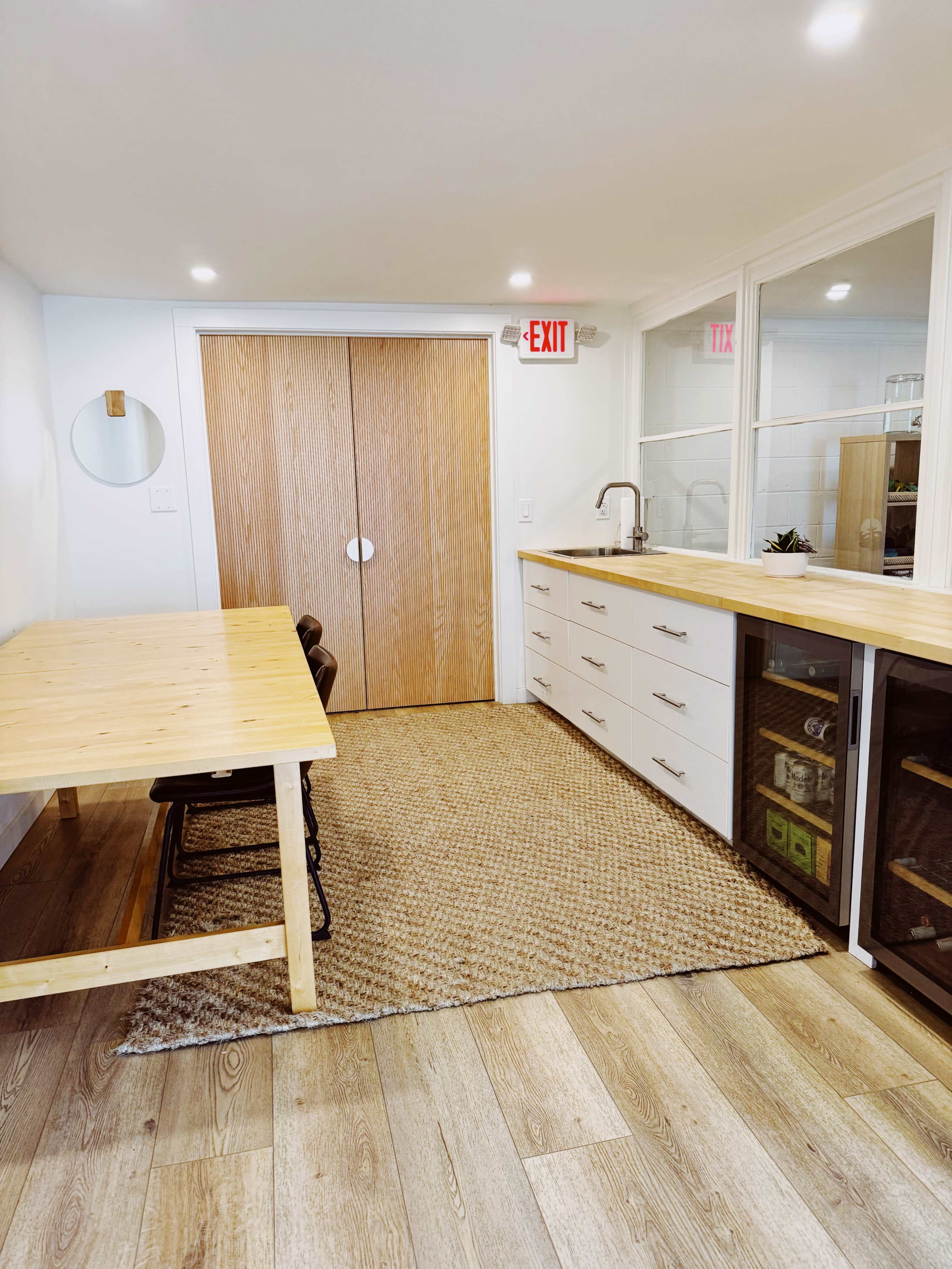 A bright, organized room featuring a wooden table with chairs, a kitchen area with cabinets, and a door leading to an exit.