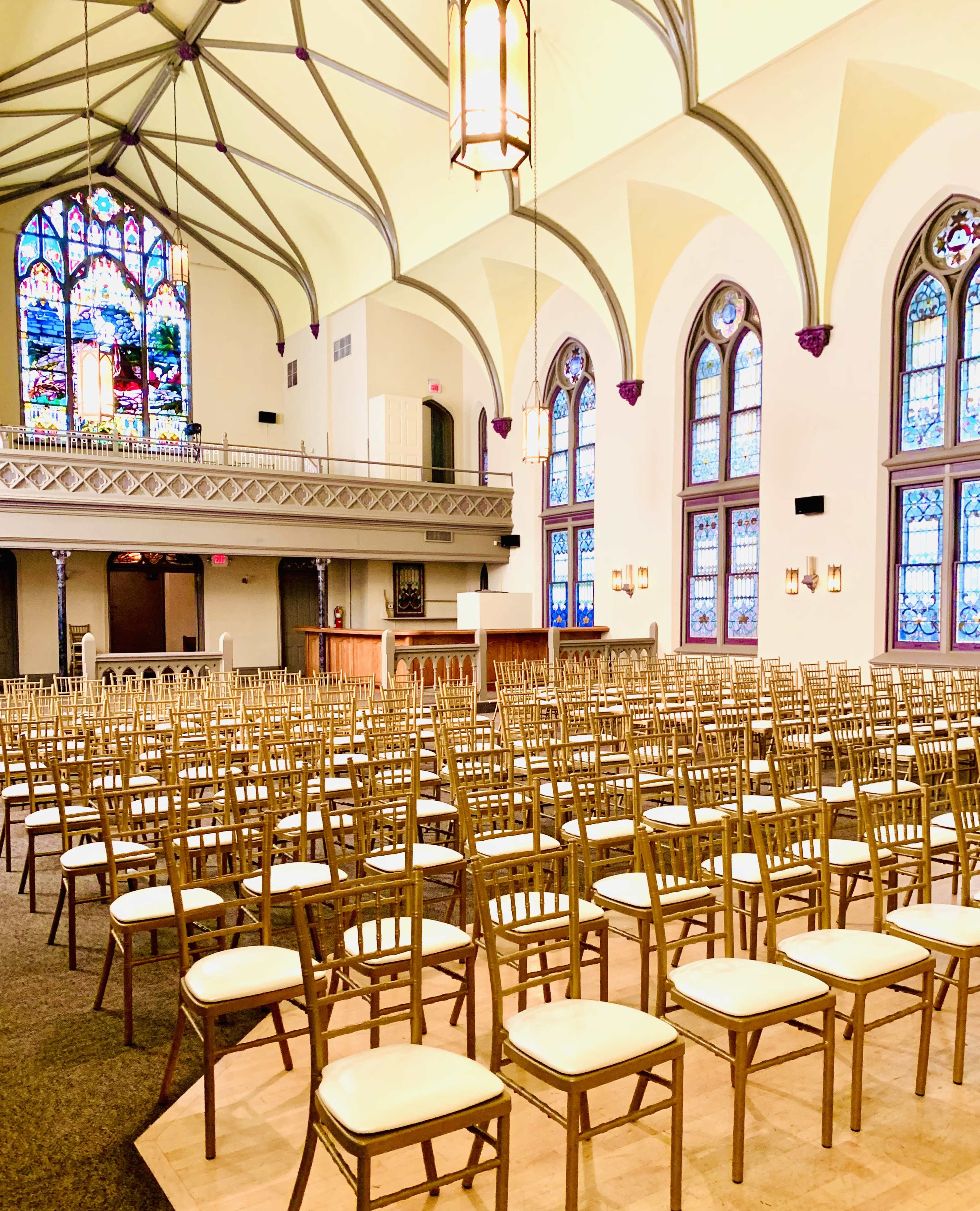 A large hall with numerous gold-colored chairs arranged in rows, featuring tall stained glass windows and an elevated stage in the background.