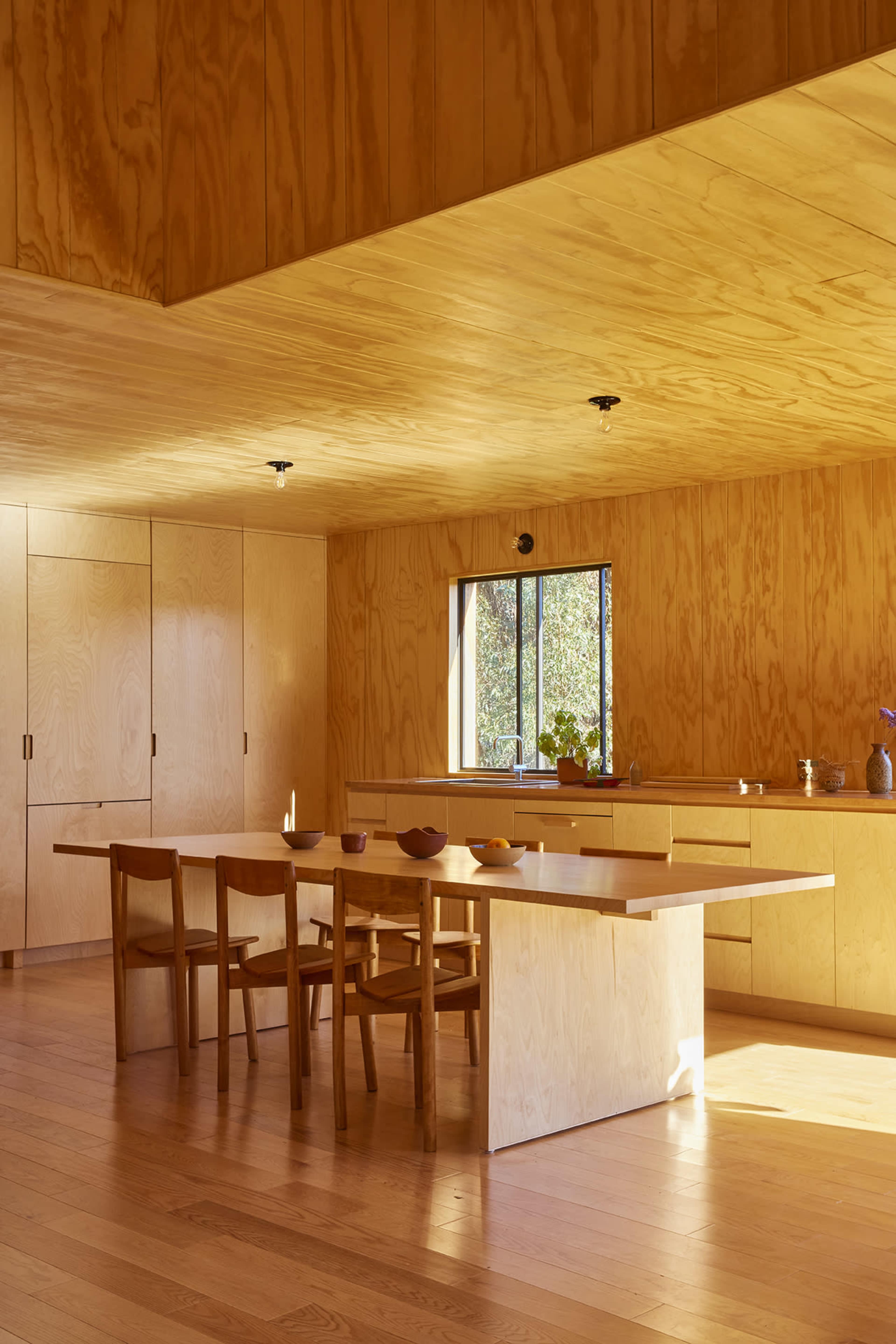 A modern, minimalist kitchen and dining area with plywood walls, a central dining table, and wooden chairs.