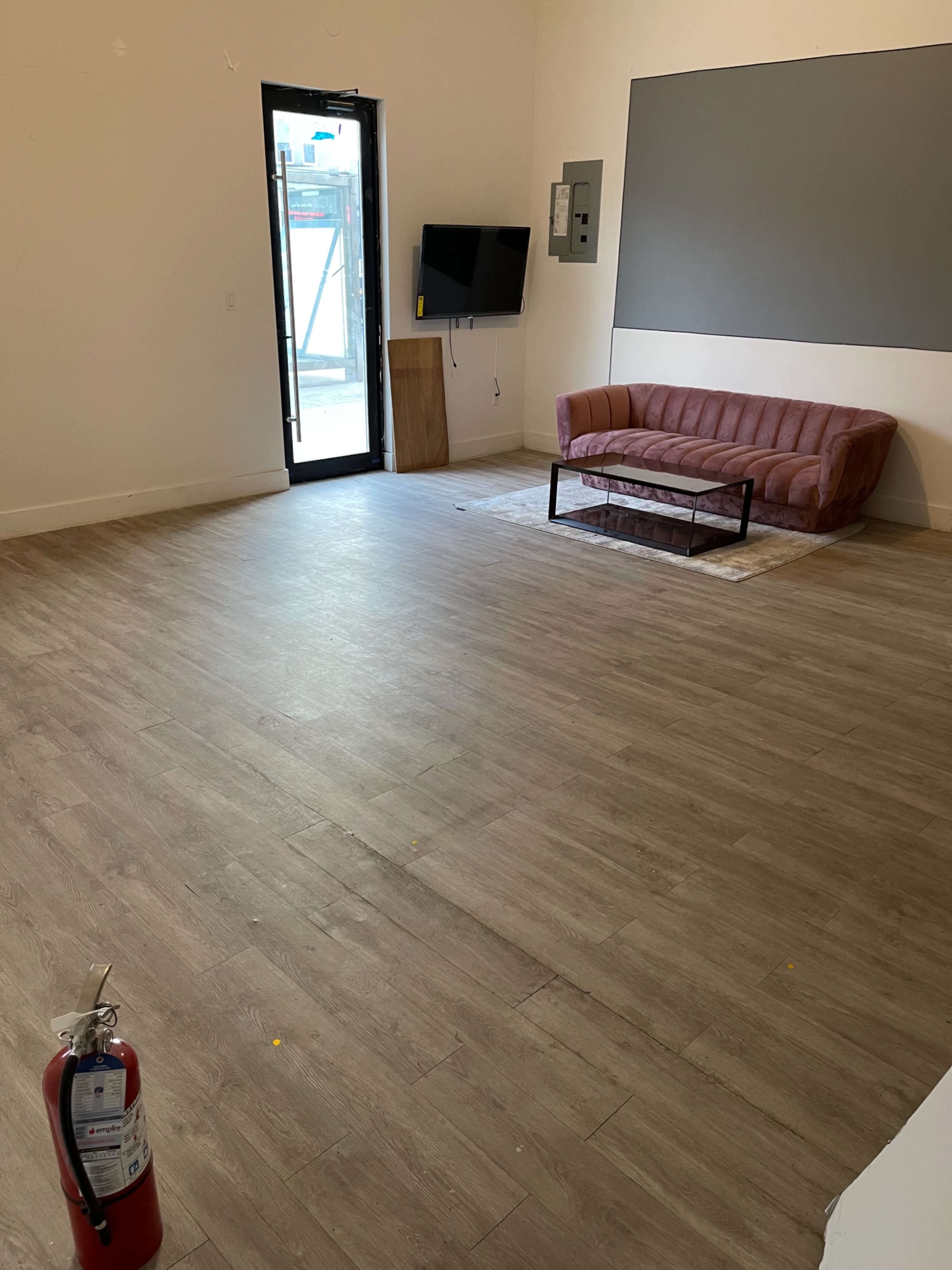 The image shows an empty room with a pink sofa, a coffee table, a wall-mounted TV, and a fire extinguisher on the floor.