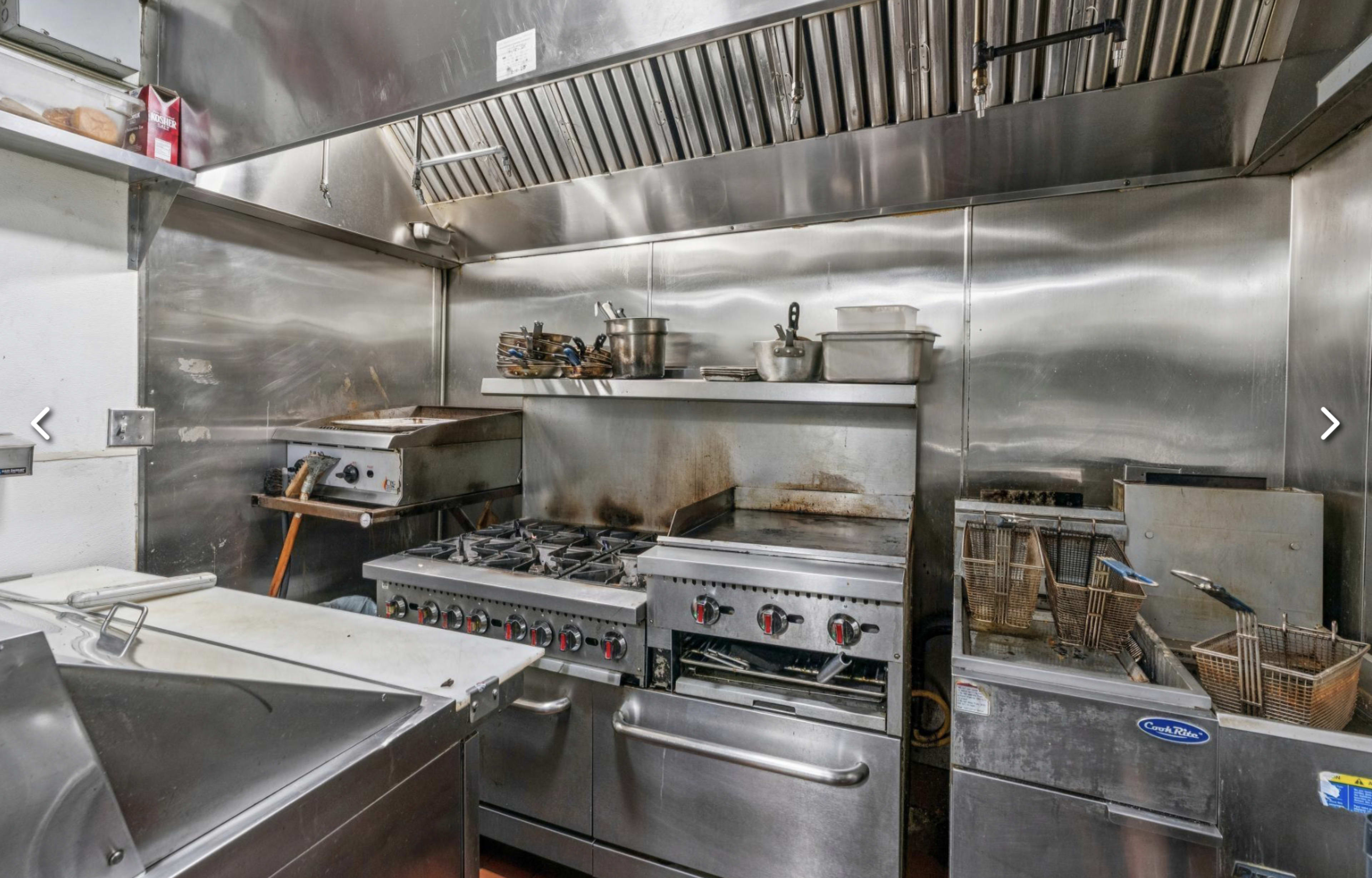 The image shows a commercial kitchen equipped with stainless steel appliances, a gas range, frying equipment, and various utensils hanging on the wall.